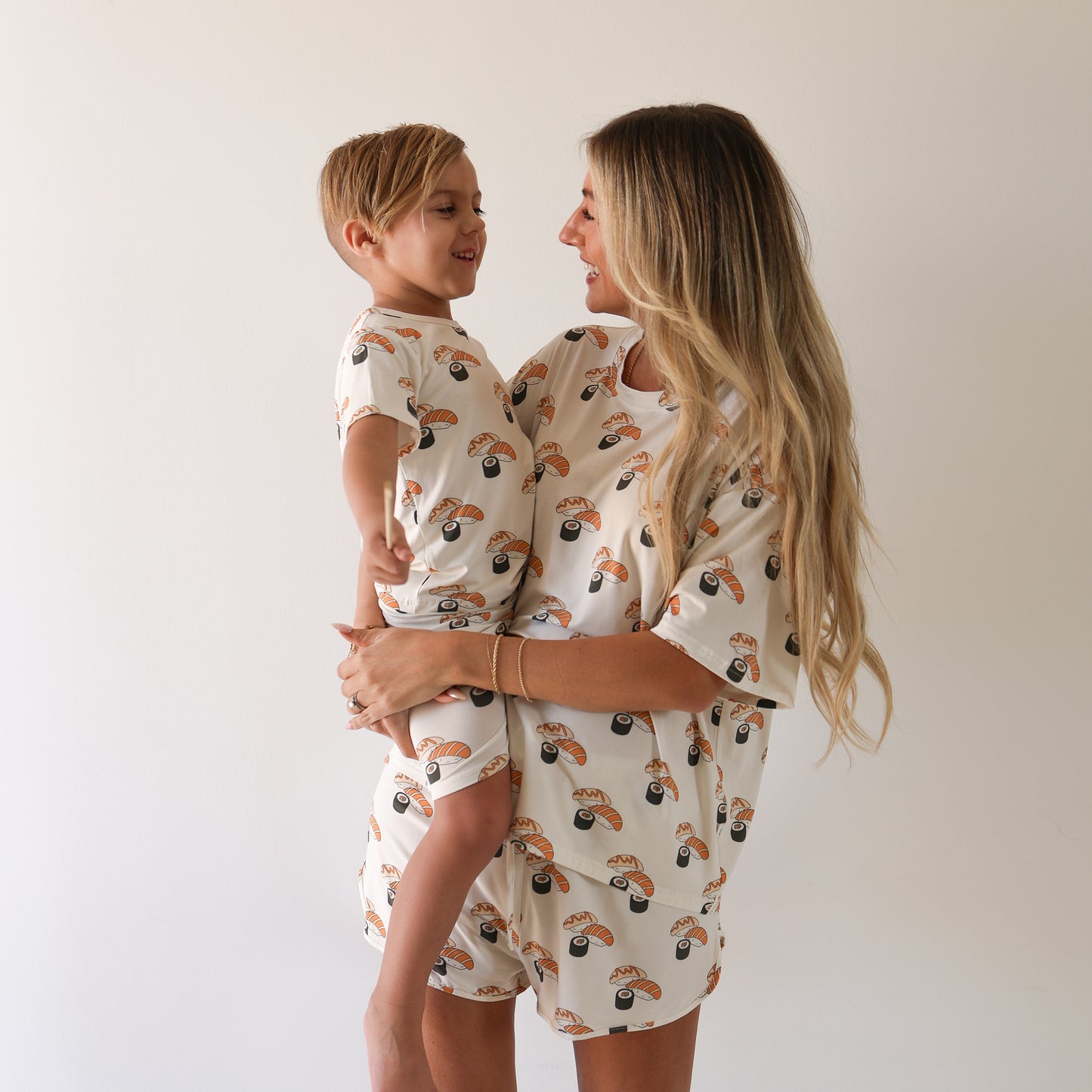 A smiling woman and young boy with long blonde hair wear matching Bamboo Short Set | Sushi pajamas from forever french baby, featuring a colorful print. Enjoy cozy, breathable bamboo sleepwear for you and your little one.