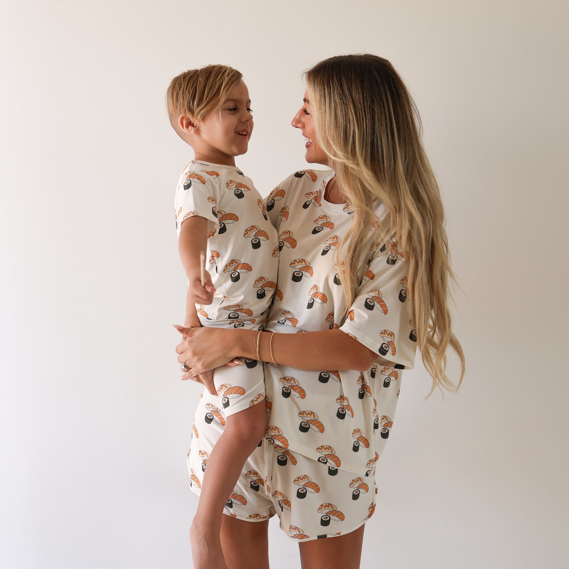 A smiling woman and young boy with long blonde hair wear matching Bamboo Short Set | Sushi pajamas from forever french baby, featuring a colorful print. Enjoy cozy, breathable bamboo sleepwear for you and your little one.
