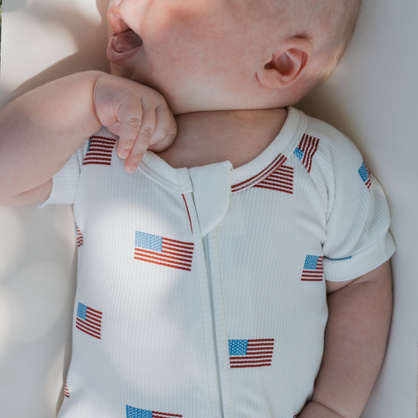 A baby in the forever french baby Shortie Bamboo Zip | Americana Ribbed onesie, featuring a white, hypoallergenic fabric with small American flags, lies on a light surface with head turned and mouth slightly open.