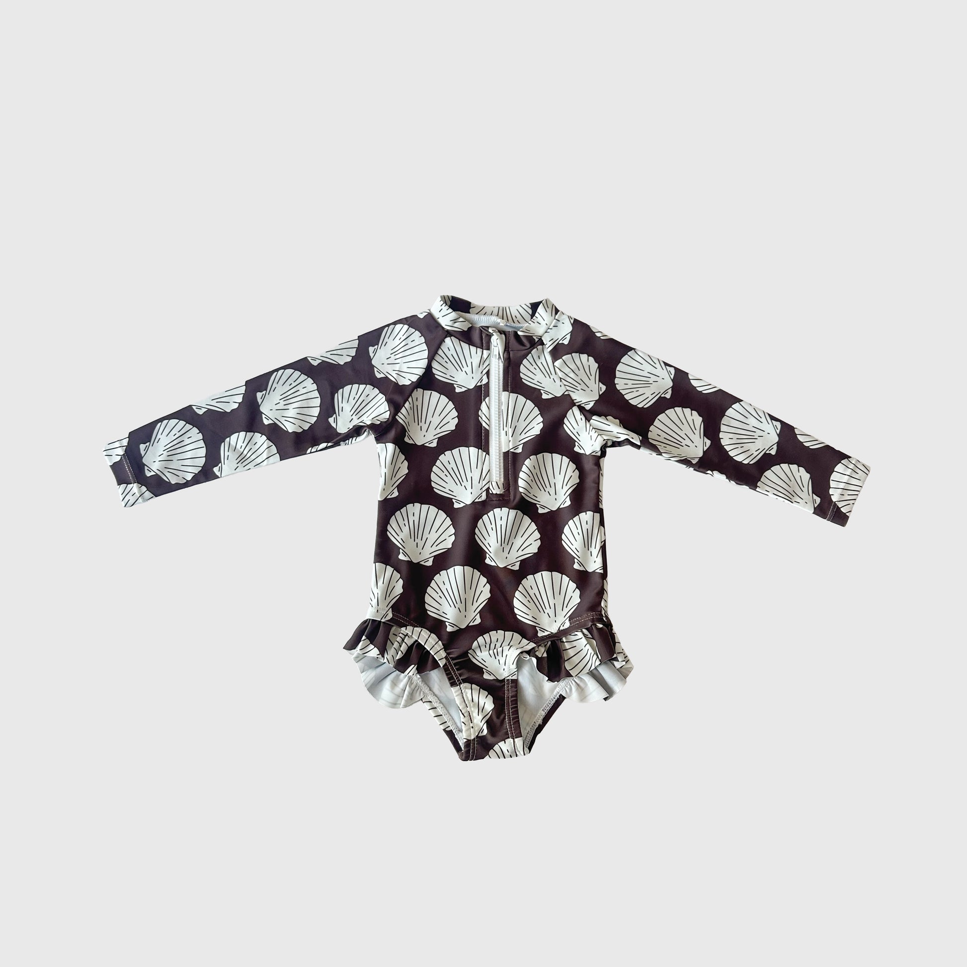 The Girl's Long Sleeve Swimsuit | Seashells by forever french baby features a dark background with white seashells, front zipper, and hip ruffles. This stylish one-piece for kids is shown on a plain light background.