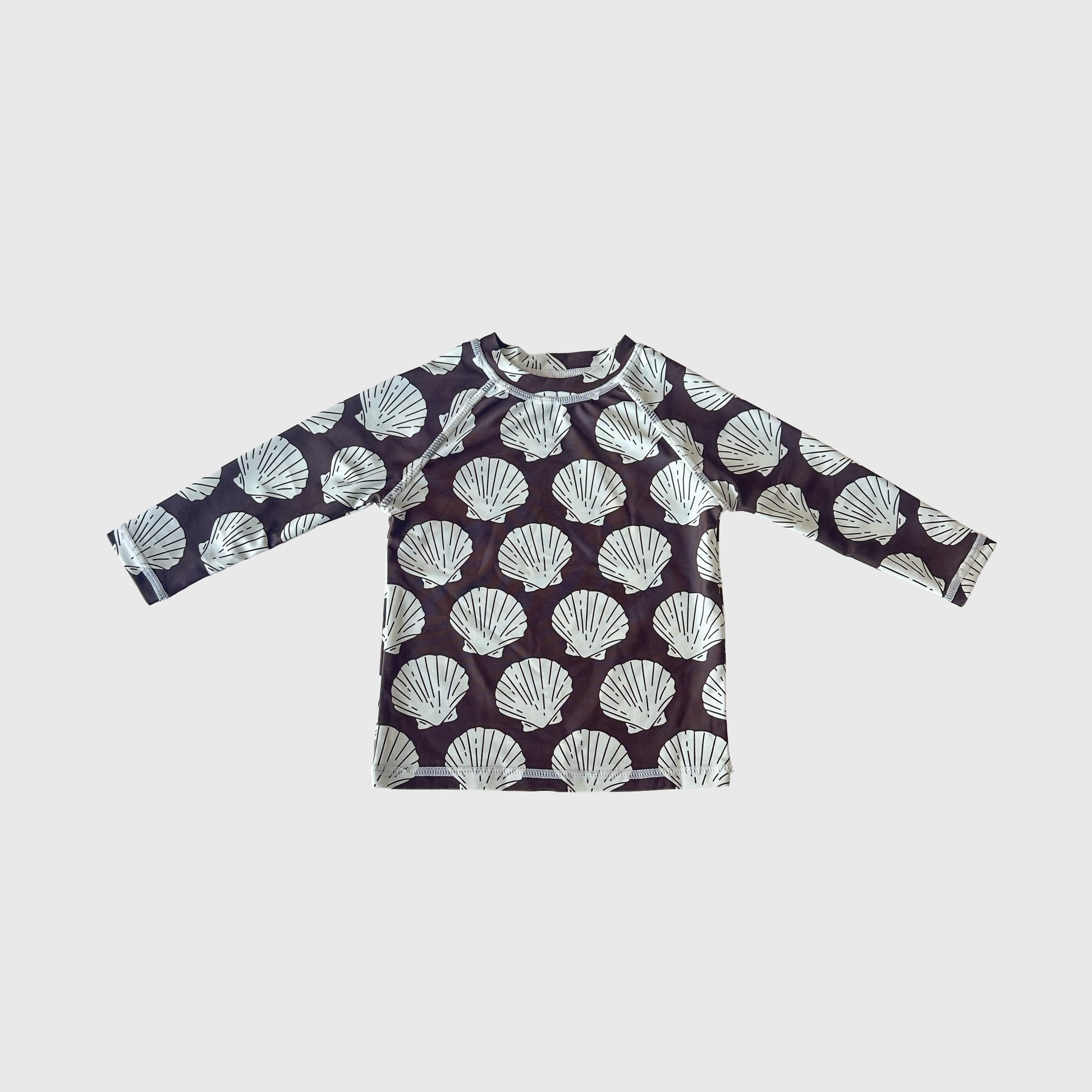The Child Rash Guard | Seashells by forever french baby features long sleeves, a dark background, and a repeating white seashell pattern—ideal for kids' swimwear or summer outfits. Displayed flat on a light gray surface.
