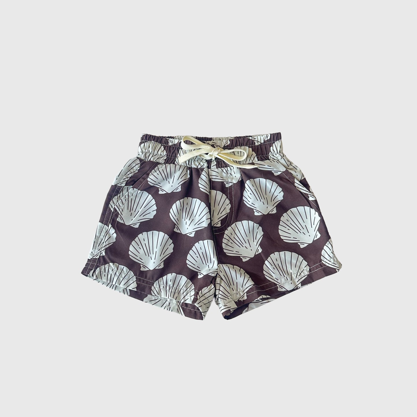 The forever french baby Child Board Short | Seashells features an elastic waistband, drawstring, and a bold seashell print on dark fabric—perfect for summer, shown on a plain light grey surface.