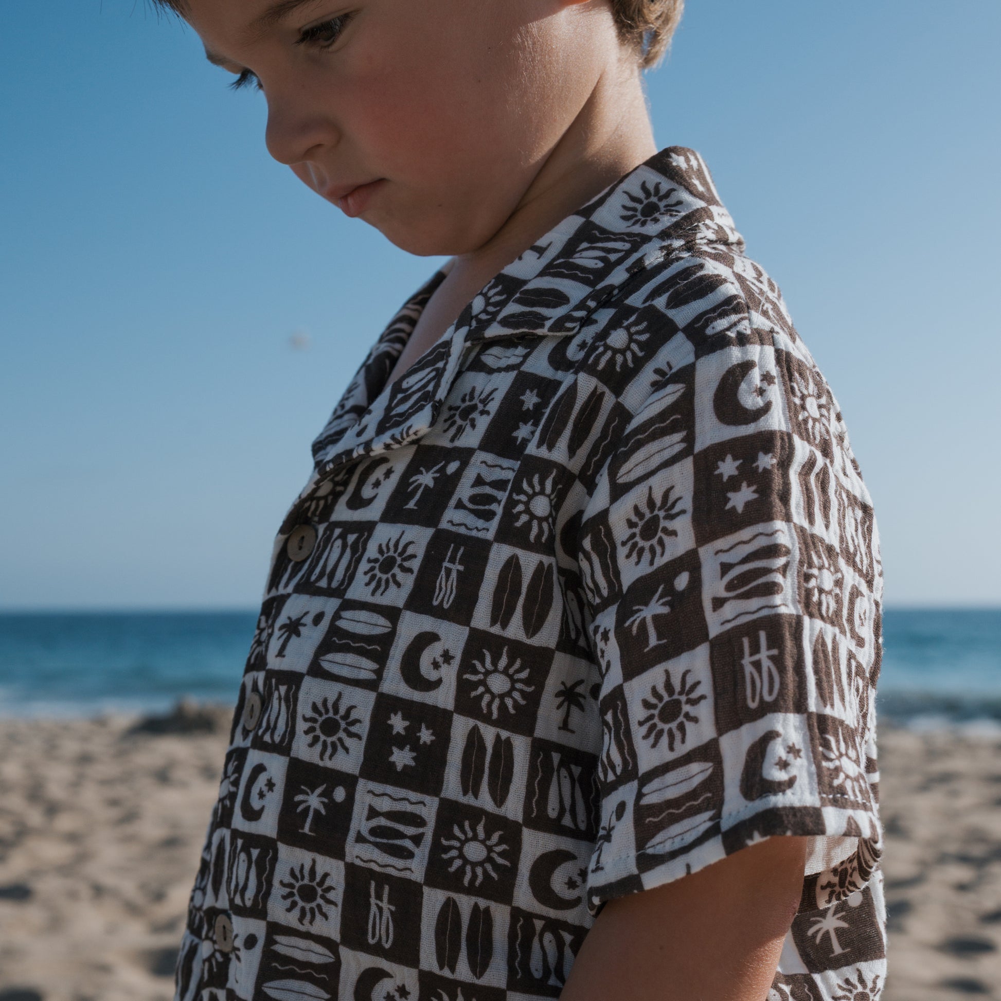 A young child wears the forever french baby Short Sleeve Muslin Button Up in Summer Check on a sandy beach, gazing down with the blue sky and ocean as a backdrop.