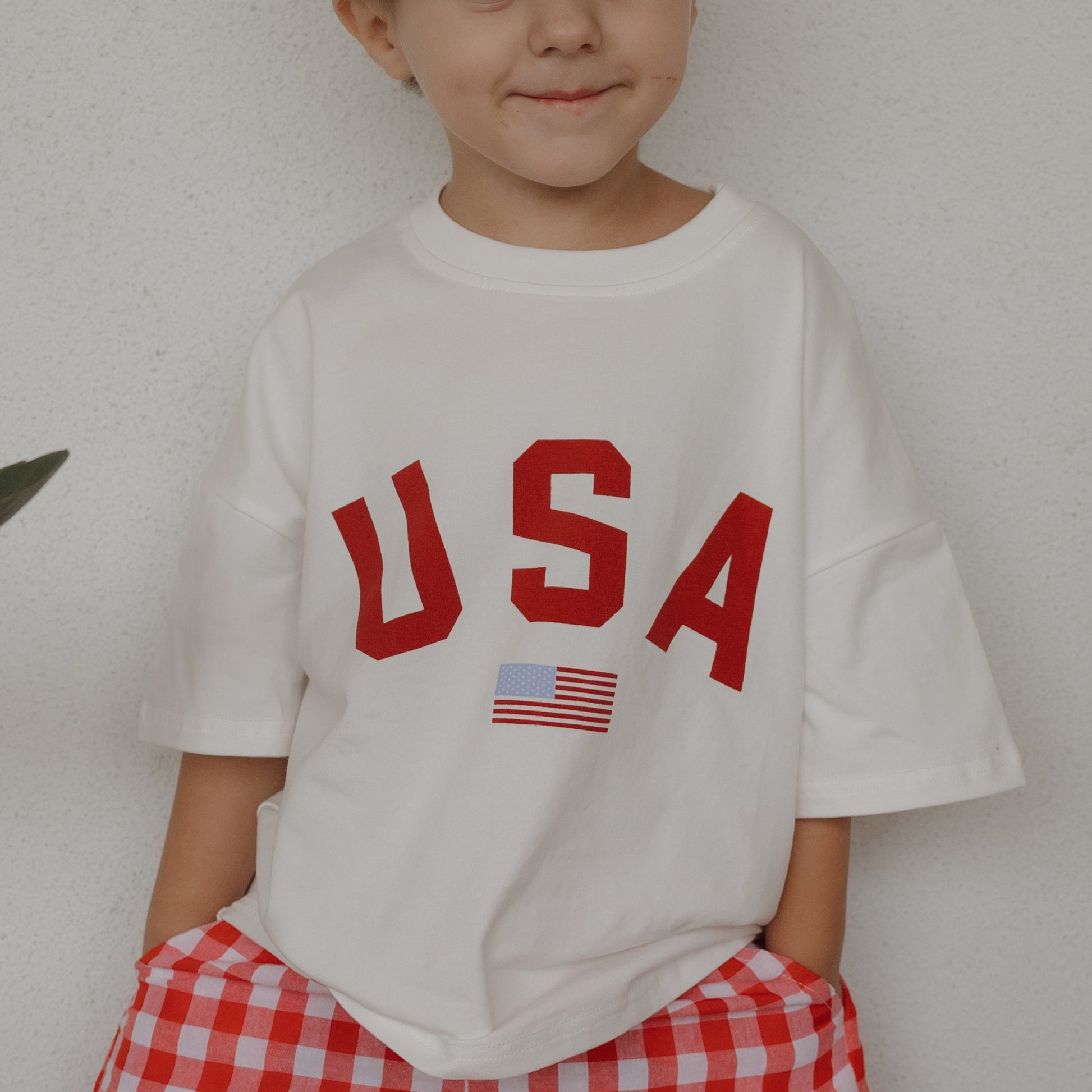 A child wears the forever french baby Child Cotton Tee | USA—100% cotton with bold red letters and a small American flag—paired with red and white checkered pants; ideal for matching family shirts on patriotic occasions.