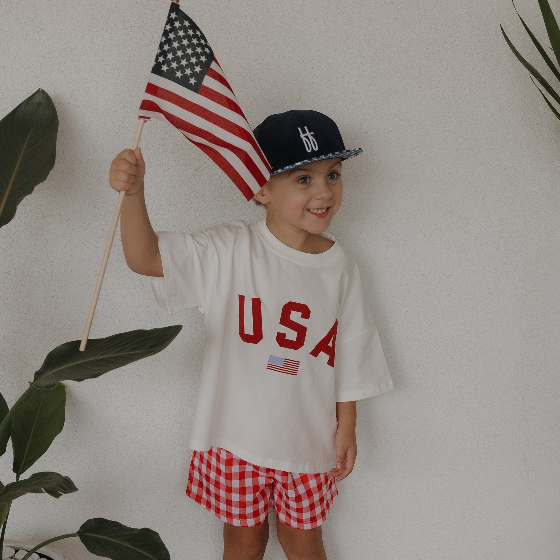 A happy child wears the forever french baby Child Cotton Tee | USA, paired with red checkered shorts and a blue cap, holding an American flag. They stand by green plants against a white wall—perfect for matching family outfits!.