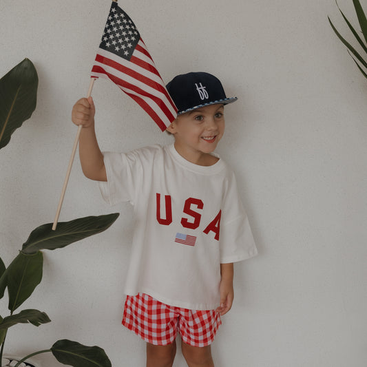 A happy child wears the forever french baby Child Cotton Tee | USA, paired with red checkered shorts and a blue cap, holding an American flag. They stand by green plants against a white wall—perfect for matching family outfits!.