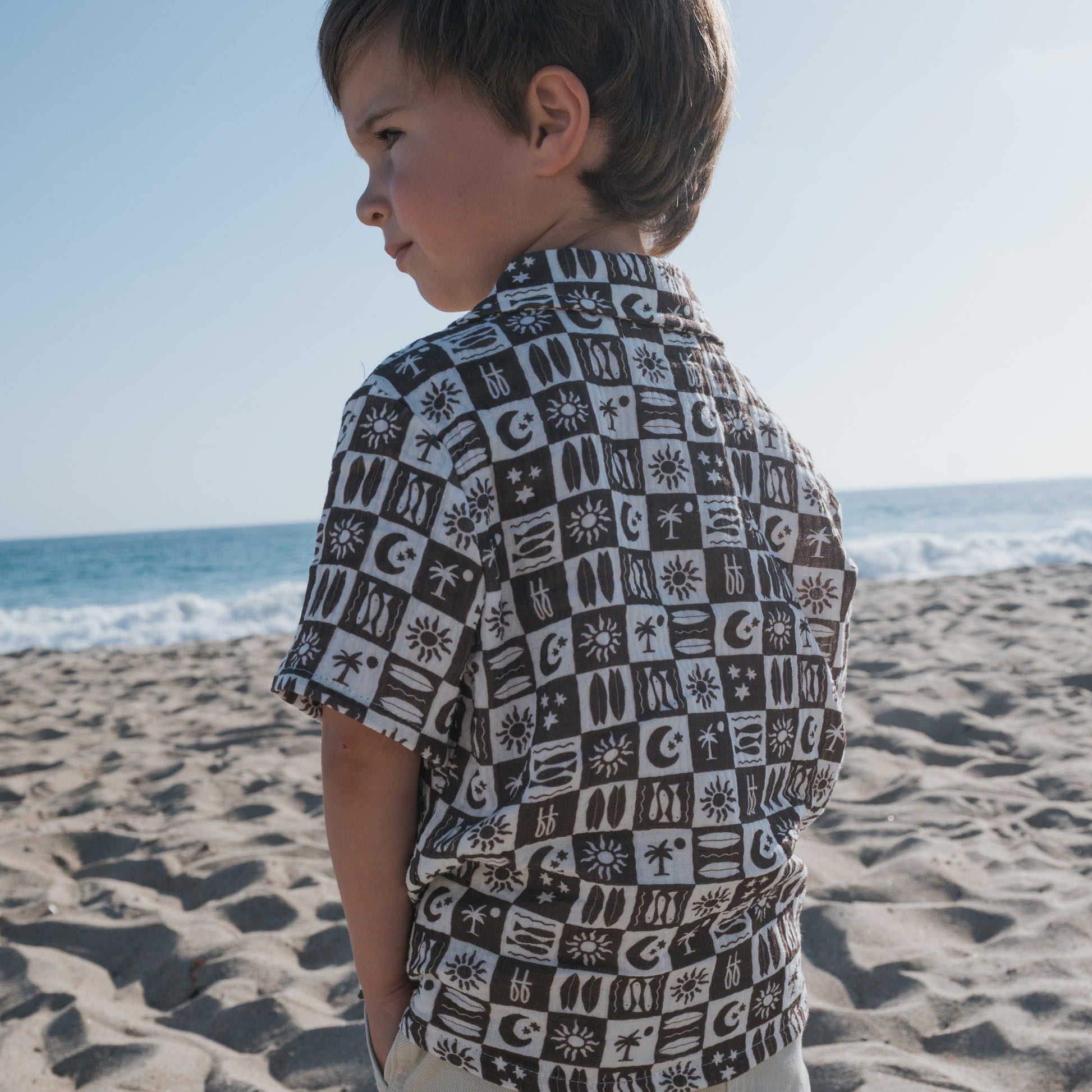 A young boy stands on a sandy beach in the forever french baby Short Sleeve Muslin Button Up | Summer Check, with the ocean and blue sky behind him.