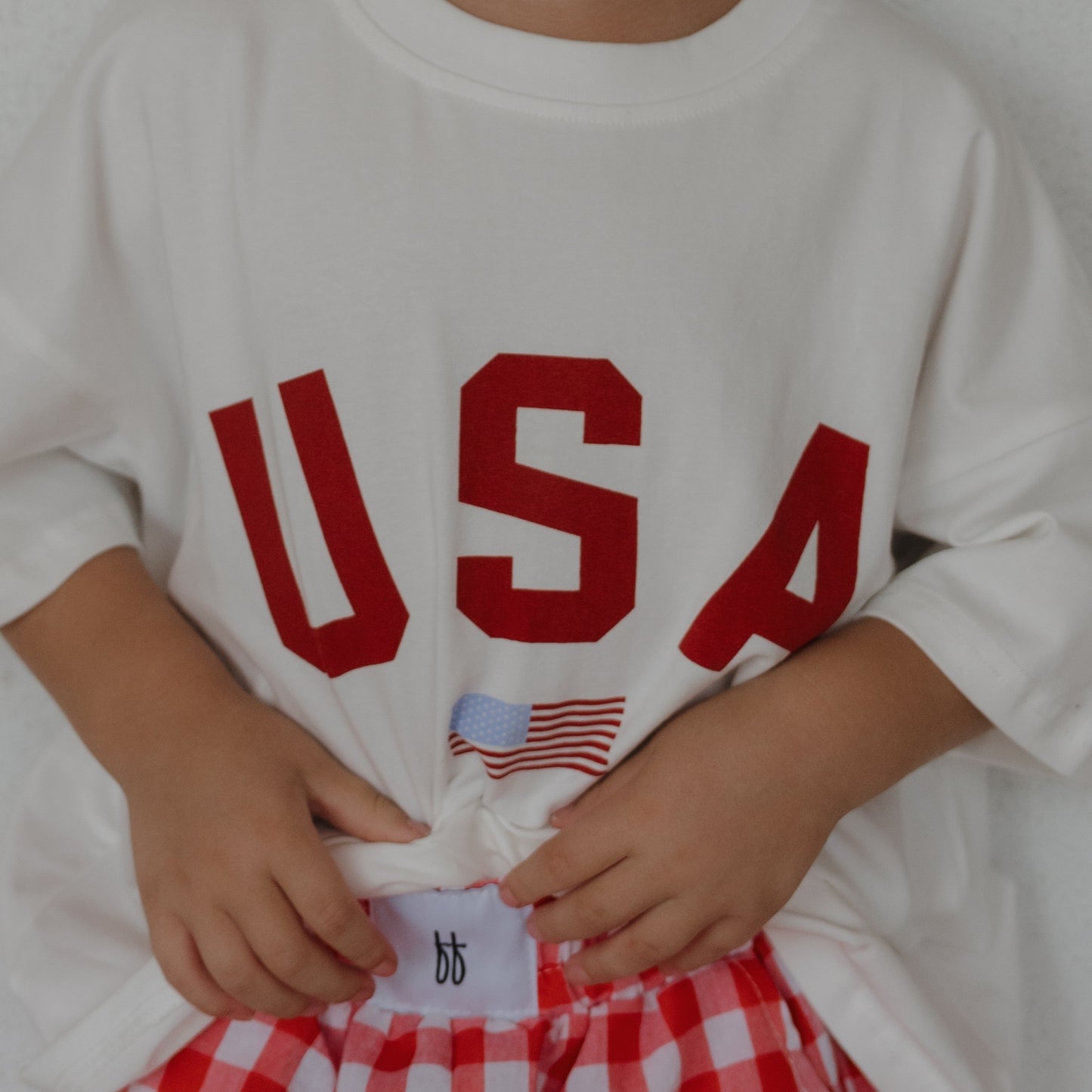 A child in the forever french baby "Child Cotton Tee | USA," featuring bold red letters and a small American flag, paired with red and white checkered shorts. The 100% cotton tee is oversized; the child's face is not visible.