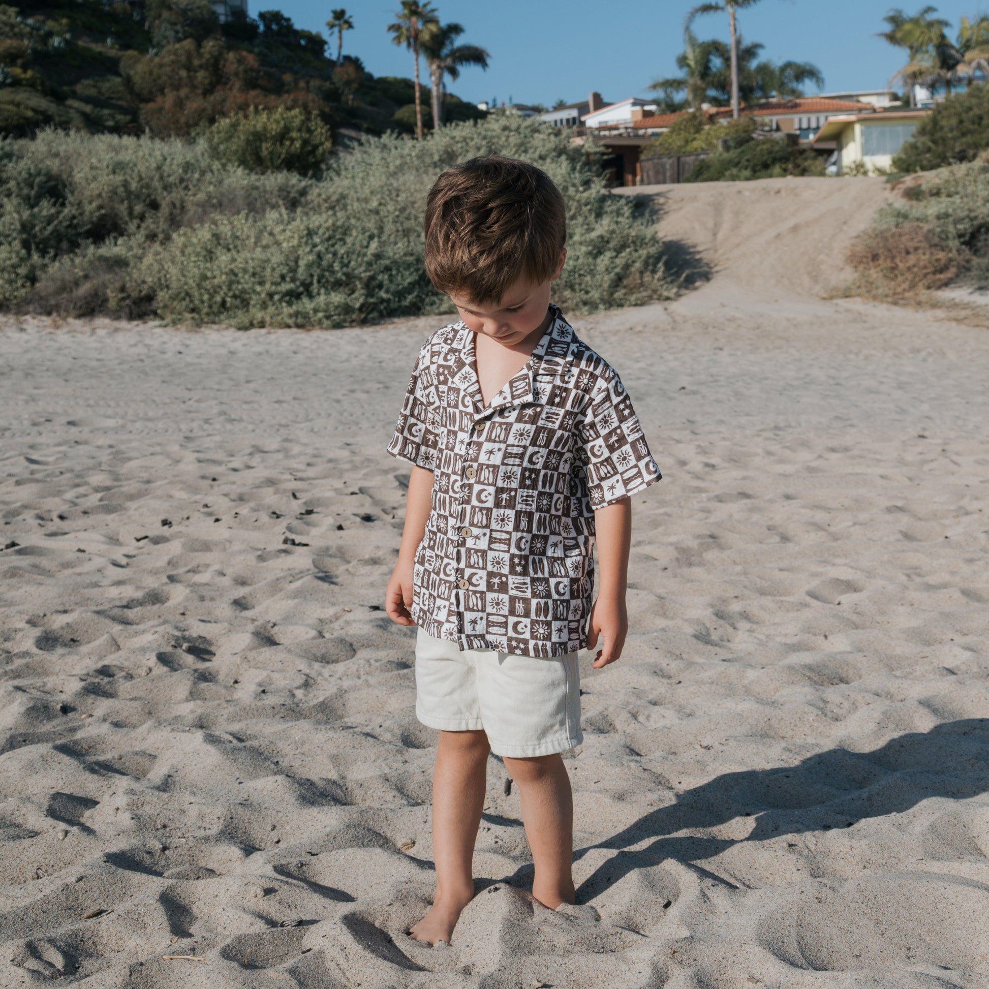 A young boy stands barefoot on a sandy beach, wearing the forever french baby Short Sleeve Muslin Button Up | Summer Check and light shorts. Green shrubs, palm trees, and houses appear behind him under a clear blue sky.