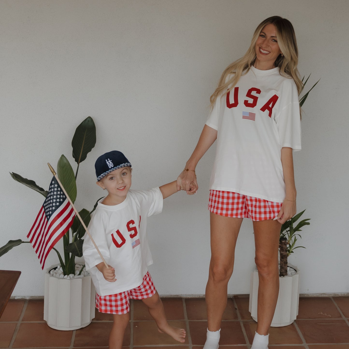 A woman and child wearing forever french baby Adult Cotton Tee | USA shirts and red checkered shorts hold hands, smiling, as the child waves an American flag. Two potted plants and a white wall are in the background.