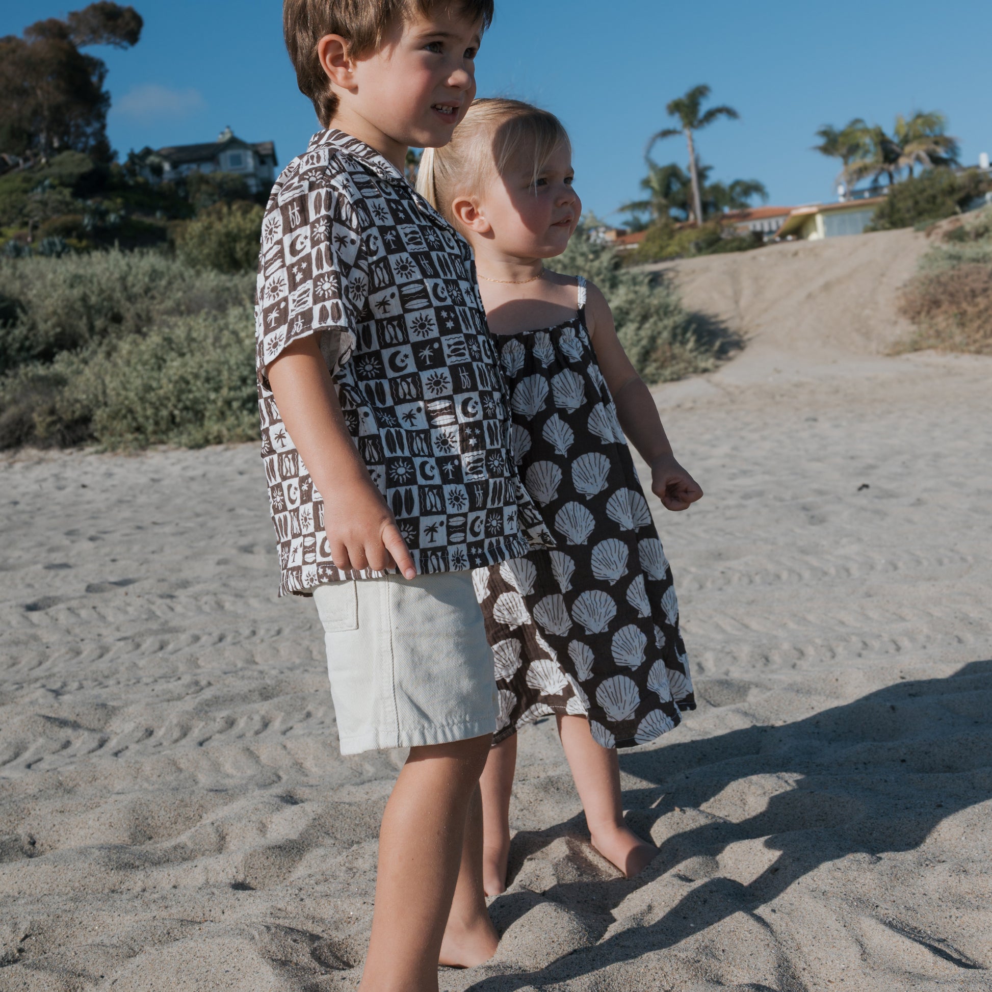 Two young children, a boy and a girl, stand barefoot on a sandy beach. The boy wears the forever french baby Short Sleeve Muslin Button Up | Summer Check and shorts; the girl wears a shell-patterned dress. Bushes, palm trees, and houses are in the background.
