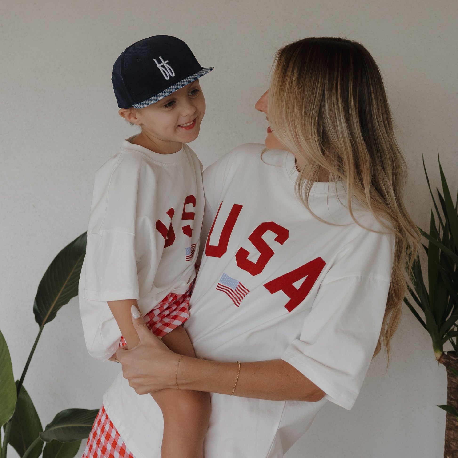 A woman and child smile at each other near green indoor plants, both wearing matching forever french baby white "Child Cotton Tee | USA" 100% cotton shirts with red checkered bottoms; the child also wears a navy blue cap.