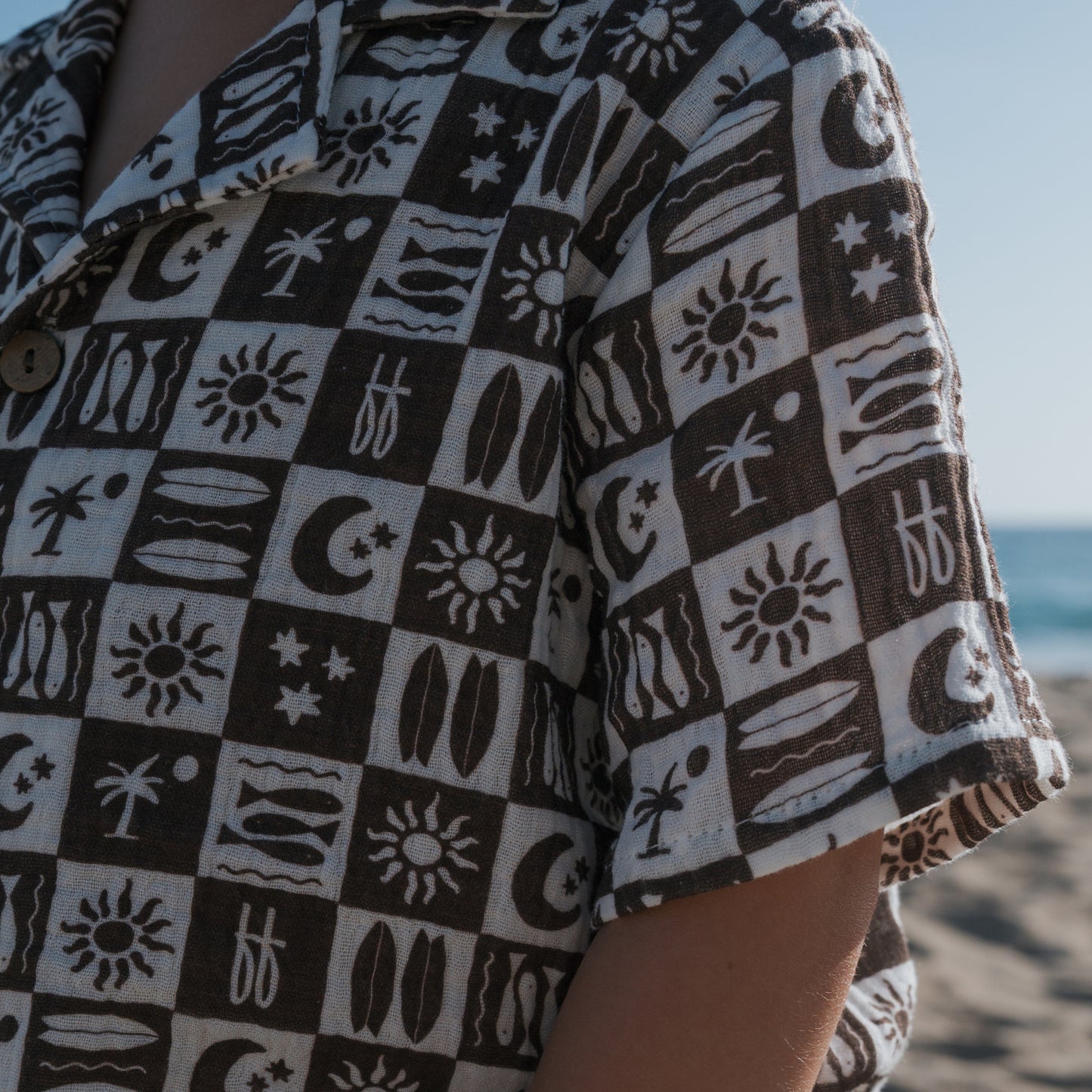 Wearing the forever french baby Short Sleeve Muslin Button Up in Summer Check, someone stands on a beach with blue sky and ocean behind them.