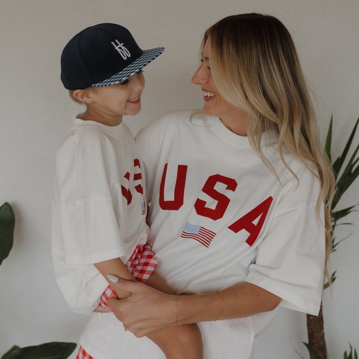 A woman and child smile at each other, both wearing forever french baby Adult Cotton Tee | USA shirts featuring flag prints. The child also wears a blue baseball cap. They stand near green plants with a plain background.