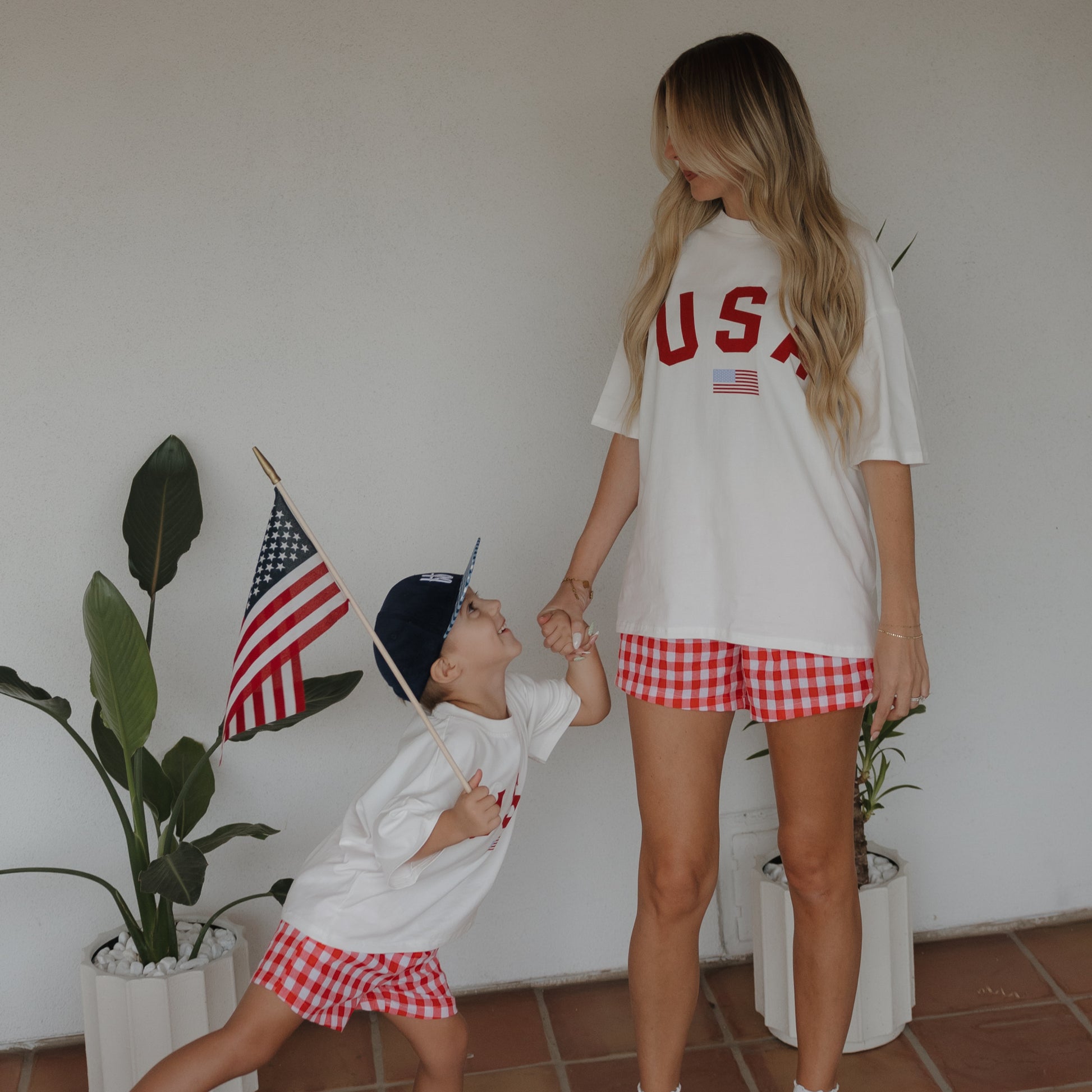 A woman and child, both in matching "USA" shirts and forever french baby's Women's Boxer Shorts | Scarlet Gingham, hold hands indoors by potted plants. The child, holding an American flag, looks up at the woman.