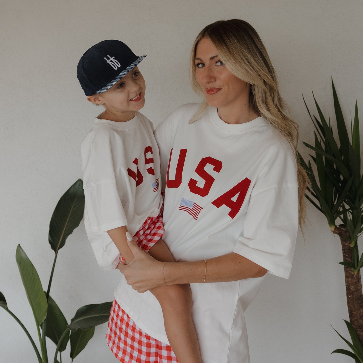 A woman and child, both in forever french baby Adult Cotton Tee | USA shirts and red-checked bottoms, smile by green plants. The child wears a navy cap as they pose together against a white wall.