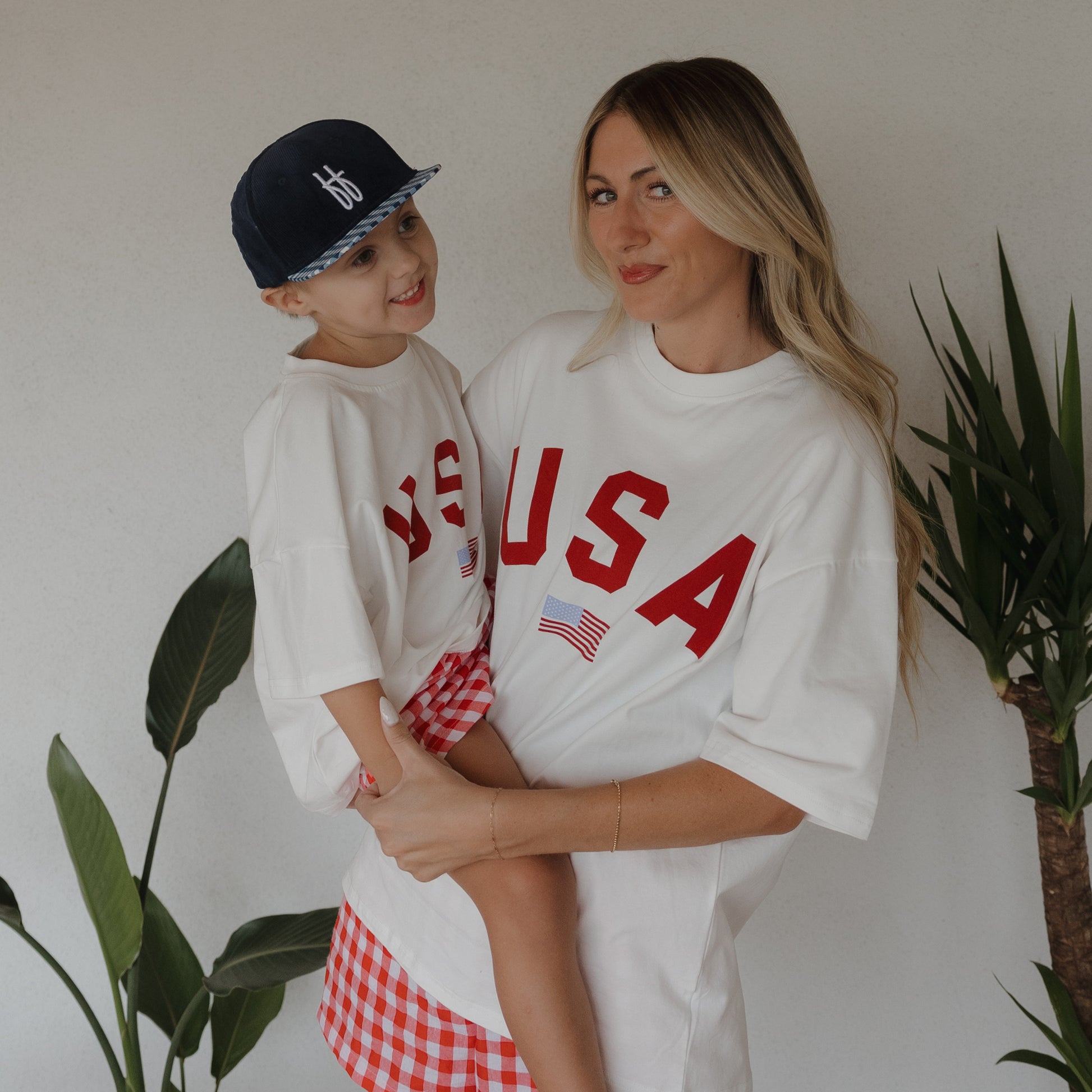 A woman and child, both in forever french baby Adult Cotton Tee | USA shirts and red-checked bottoms, smile by green plants. The child wears a navy cap as they pose together against a white wall.