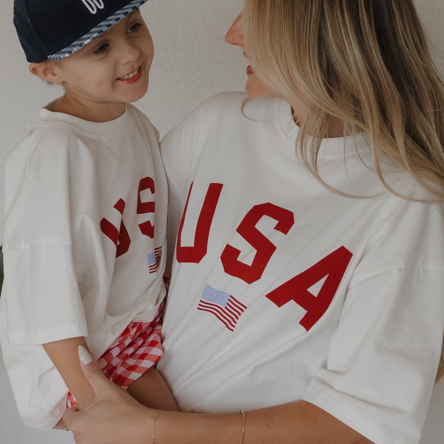 A woman wears the forever french baby Adult Cotton Tee | USA, featuring "USA" and a small flag. Her smiling child matches in an oversized version, blue cap, and red checkered shorts. Both tees are 100% cotton and unisex.