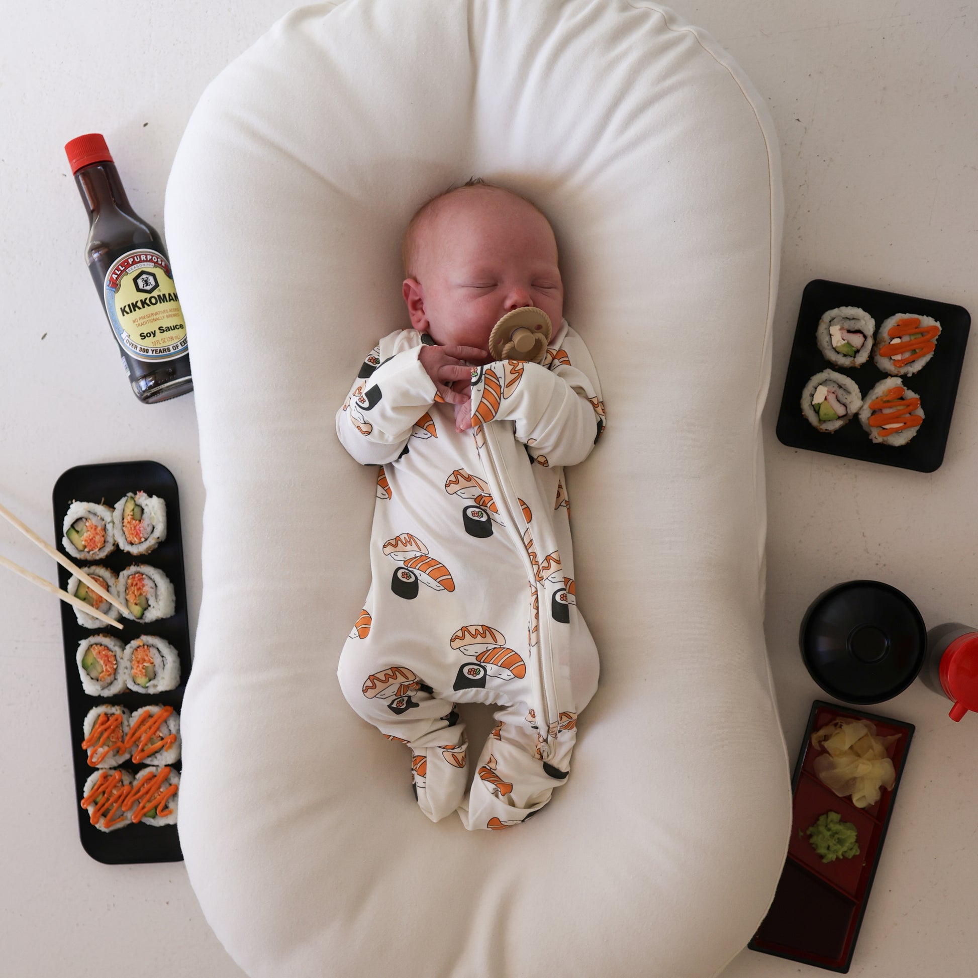 A baby in forever french baby Bamboo Zip Pajamas | Sushi sleeps on a white cushion, surrounded by sushi plates, soy sauce, chopsticks, and condiments against a white background.