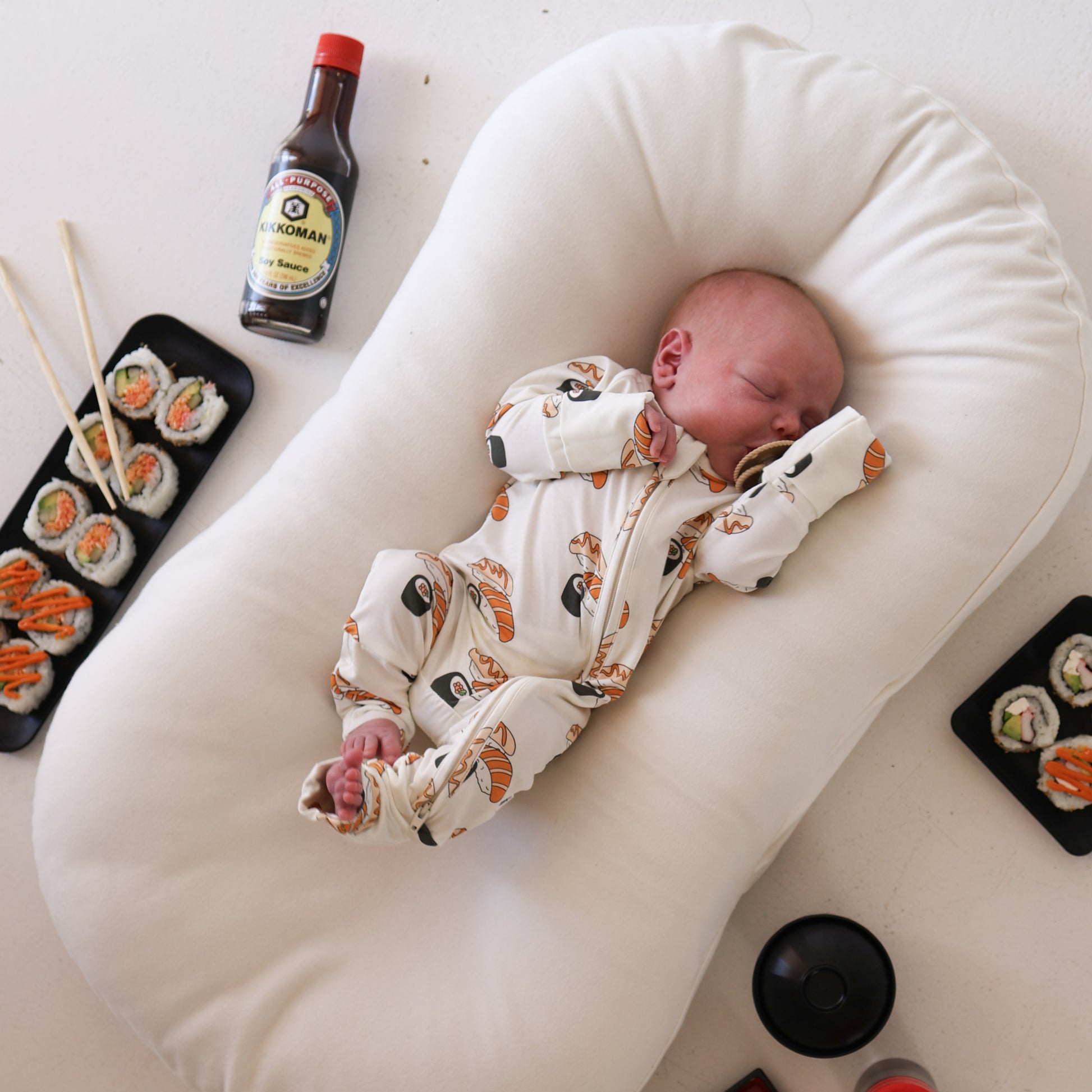 A baby wearing forever french baby's Bamboo Zip Pajamas | Sushi print naps on a white cushion, surrounded by trays of sushi, a soy sauce bottle, and chopsticks on a white surface.