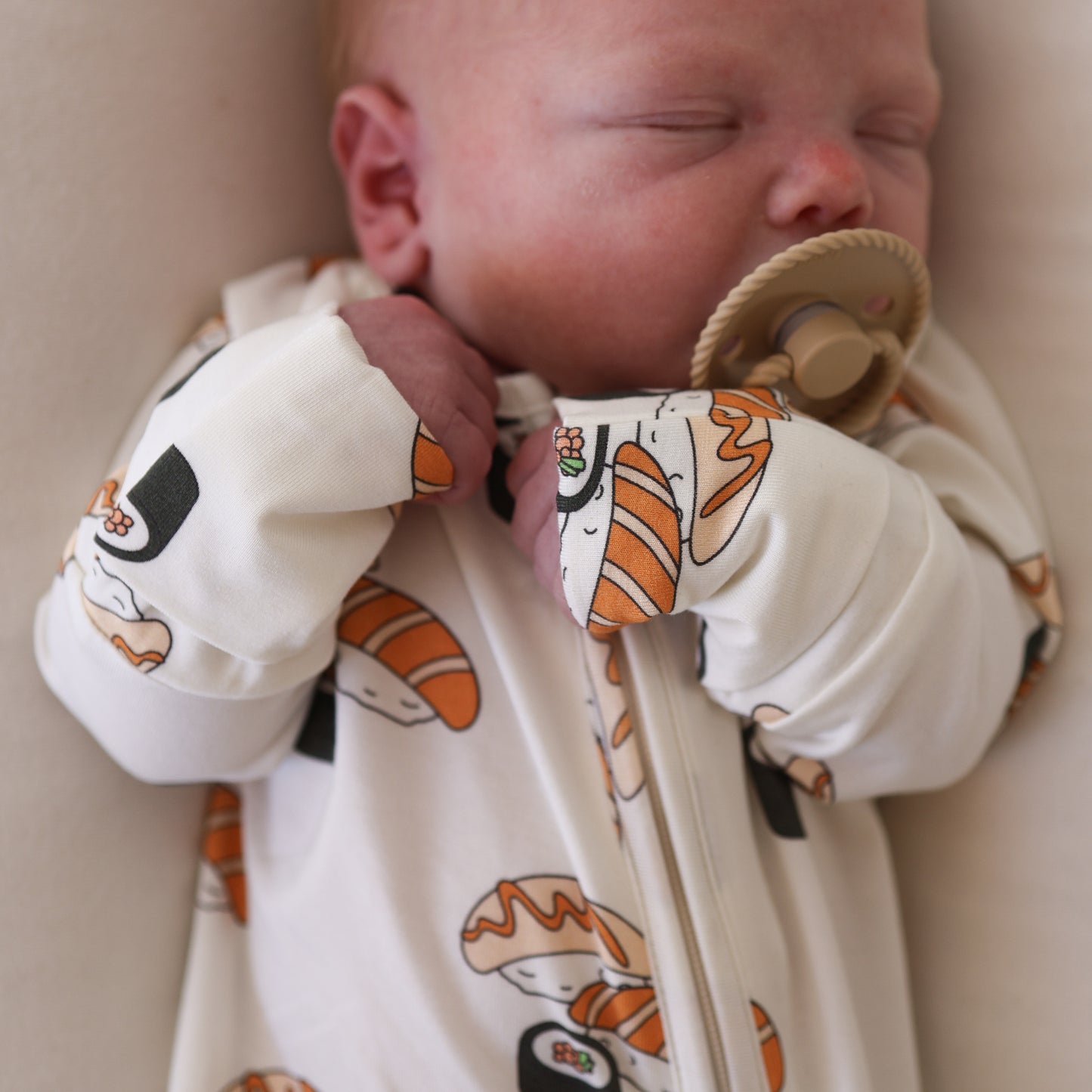 A sleeping baby, dressed in forever french baby Bamboo Zip Pajamas | Sushi—white with colorful sushi patterns—holds a tan pacifier near their mouth and rests peacefully.