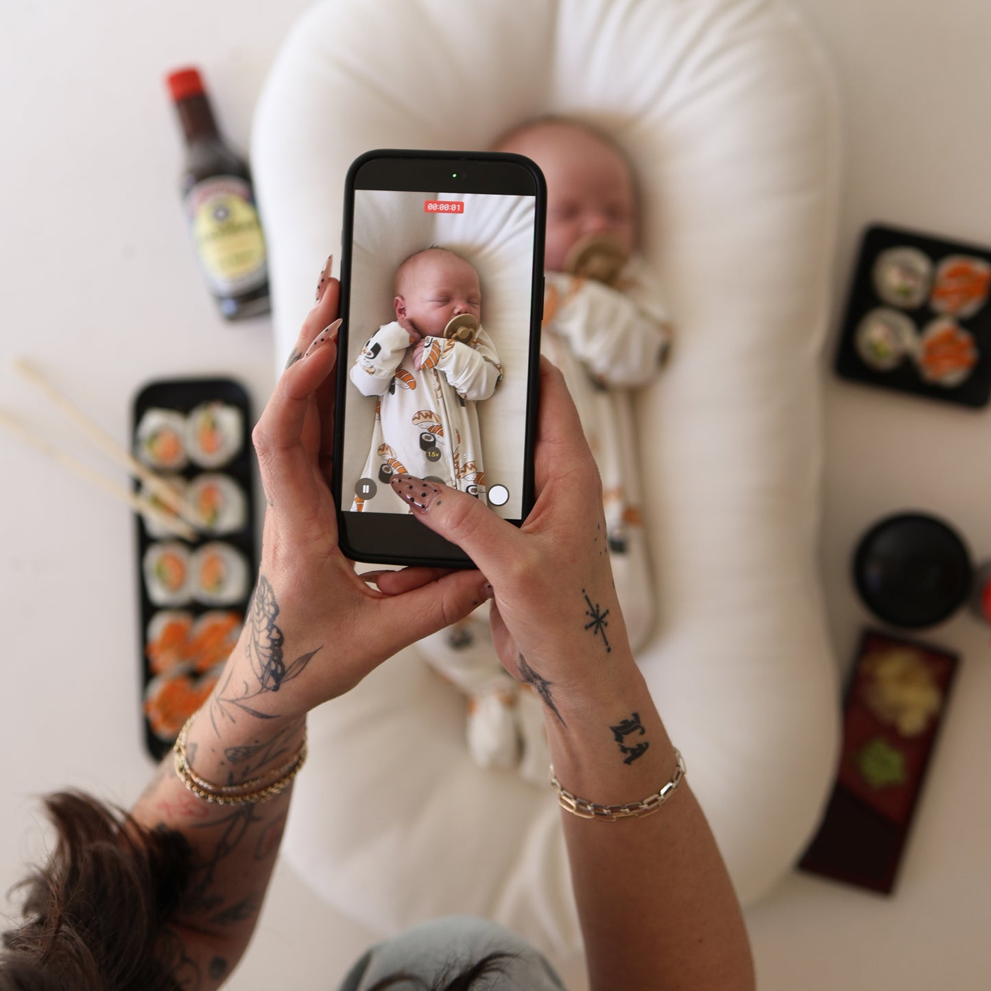 A tattooed person snaps a photo of a sleeping baby on a white cushion, wearing forever french baby's Bamboo Zip Pajamas | Sushi, surrounded by sushi, chopsticks, and soy sauce.