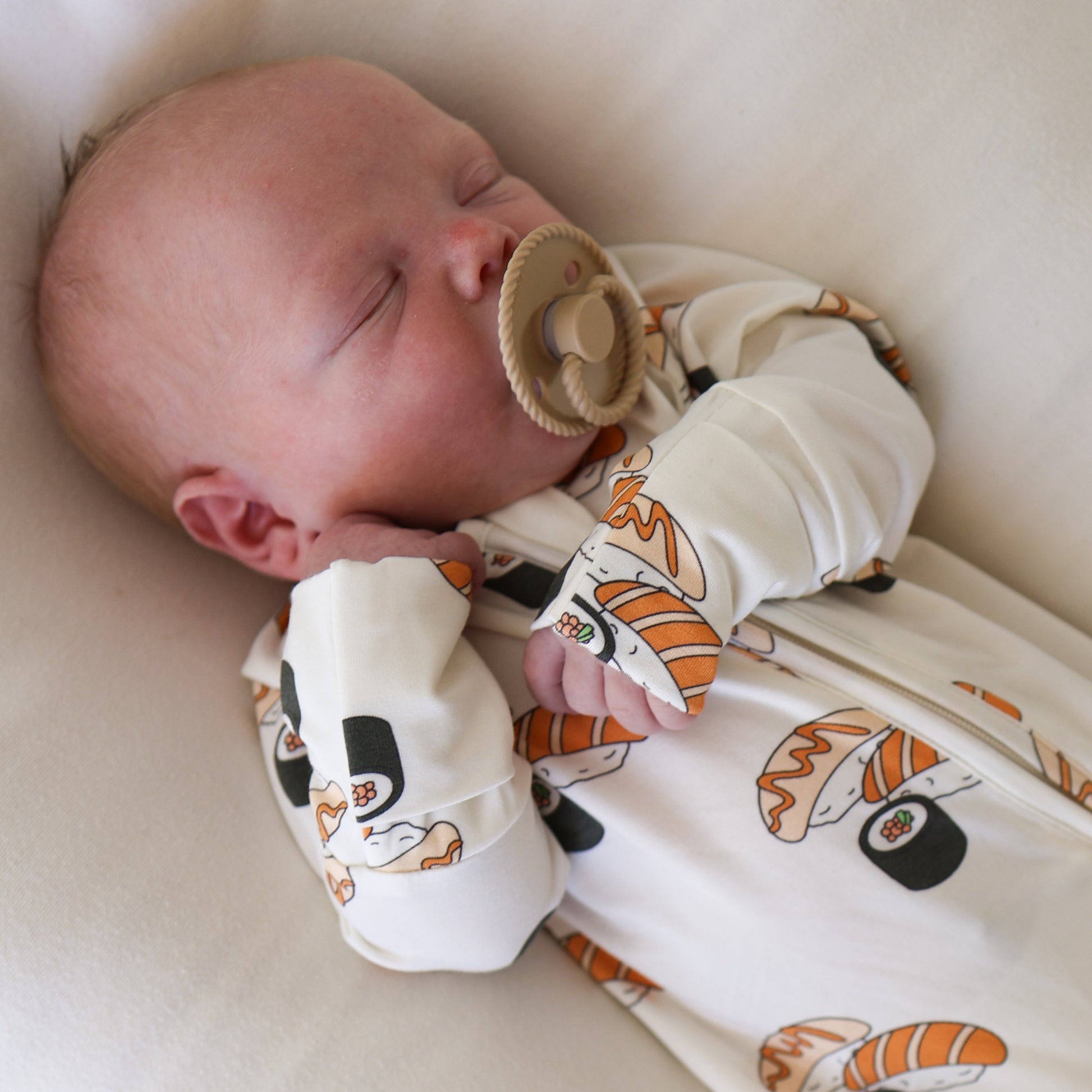 A sleeping baby with a pacifier, dressed in forever french baby Bamboo Zip Pajamas | Sushi—a hypoallergenic white onesie with sushi designs—rests on a soft, light-colored surface.
