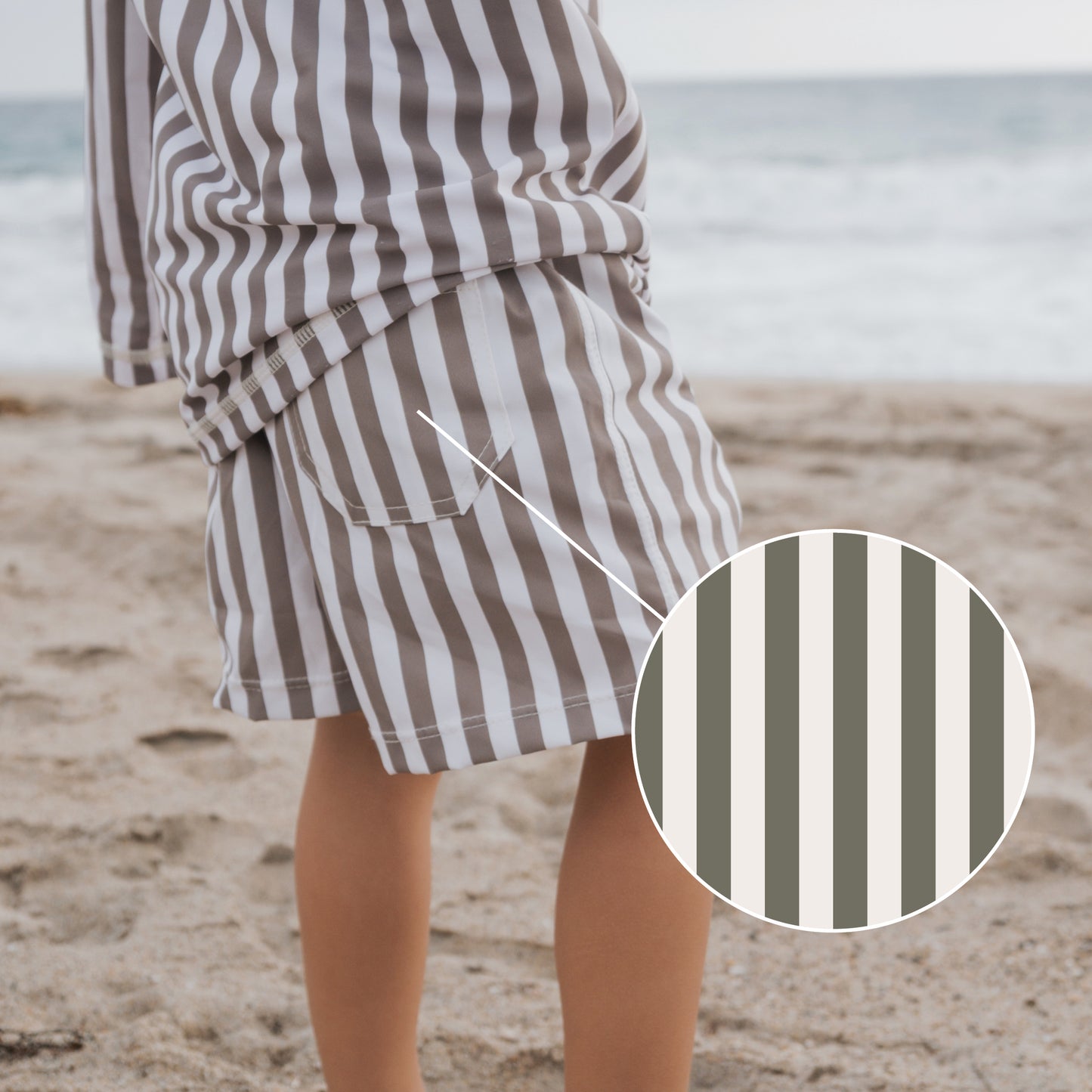 A child stands on a sandy beach in forever french baby’s Olive Stripe Child Board Short, with a circular zoom-in highlighting the olive and white stripes. The ocean is visible in the background, evoking a classic summer style.