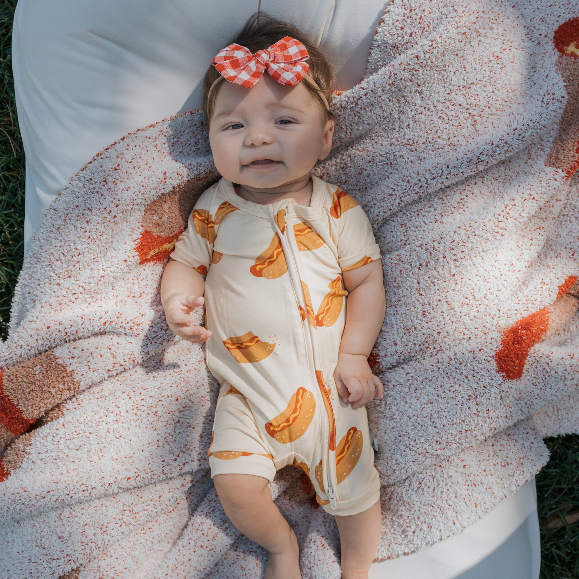 A baby wearing the forever french baby Shortie Bamboo Zip | Hot Dogs romper lies on a patterned blanket outdoors, smiling at the camera with a red-and-white checkered bow headband.
