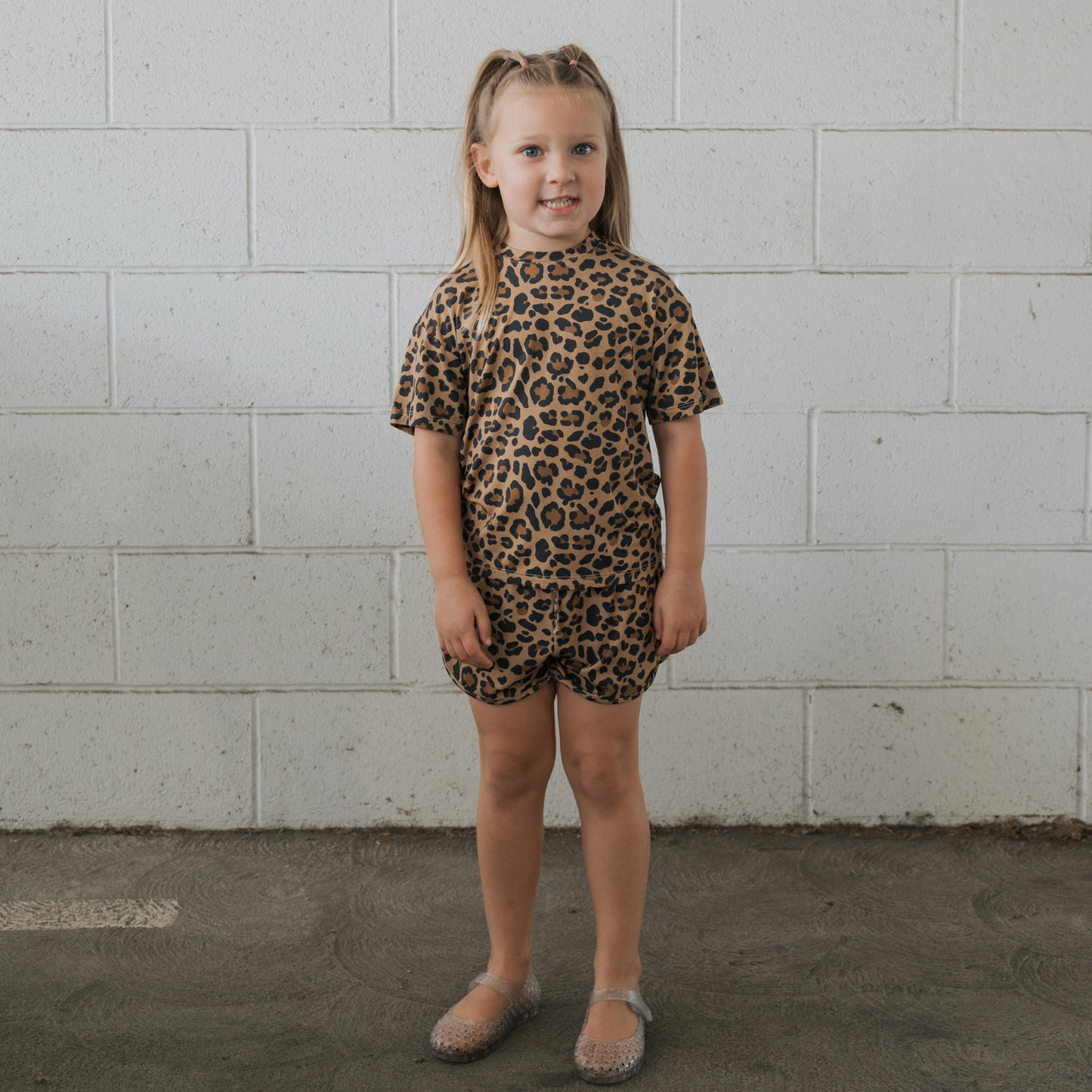 A young girl stands in front of a white brick wall, wearing the forever french baby Girl's Bamboo Short Set | Leopard—breathable bamboo T-shirt and shorts—with clear plastic shoes. She faces forward and smiles slightly.