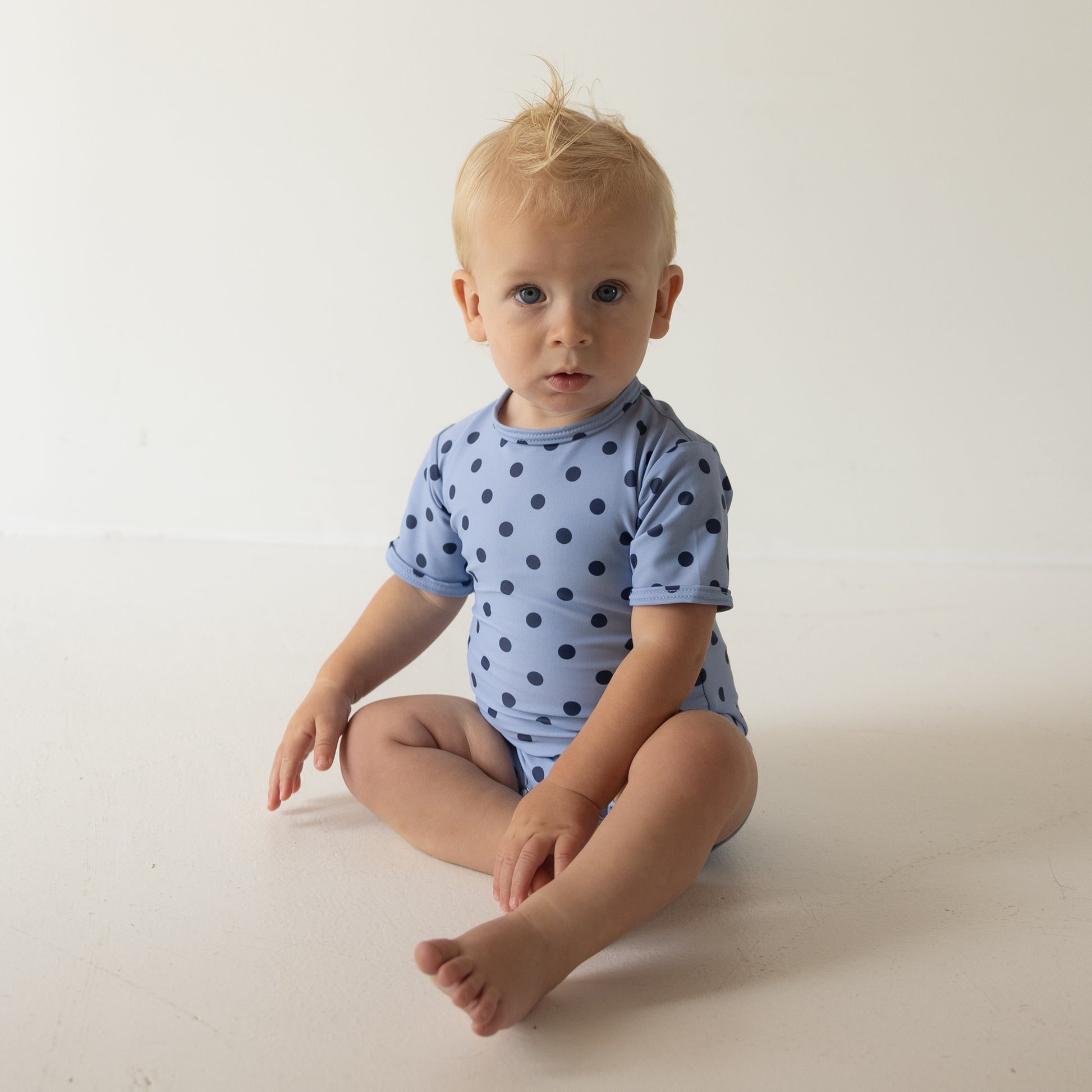 A blonde baby sits on the floor in a light blue forever french baby Child Shortie Swimsuit | Sully with dark blue polka dots, looking at the camera against a plain white background.