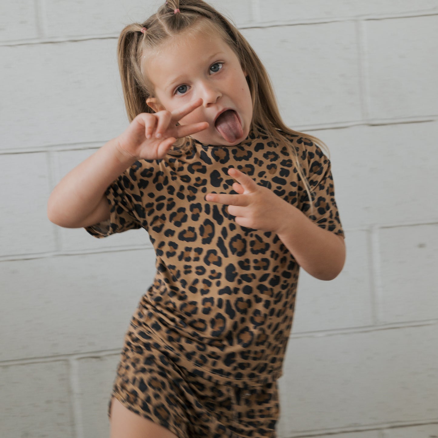 A young girl wears the forever french baby Girl's Bamboo Short Set | Leopard, standing in front of a white brick wall, smiling and flashing peace signs with both hands.