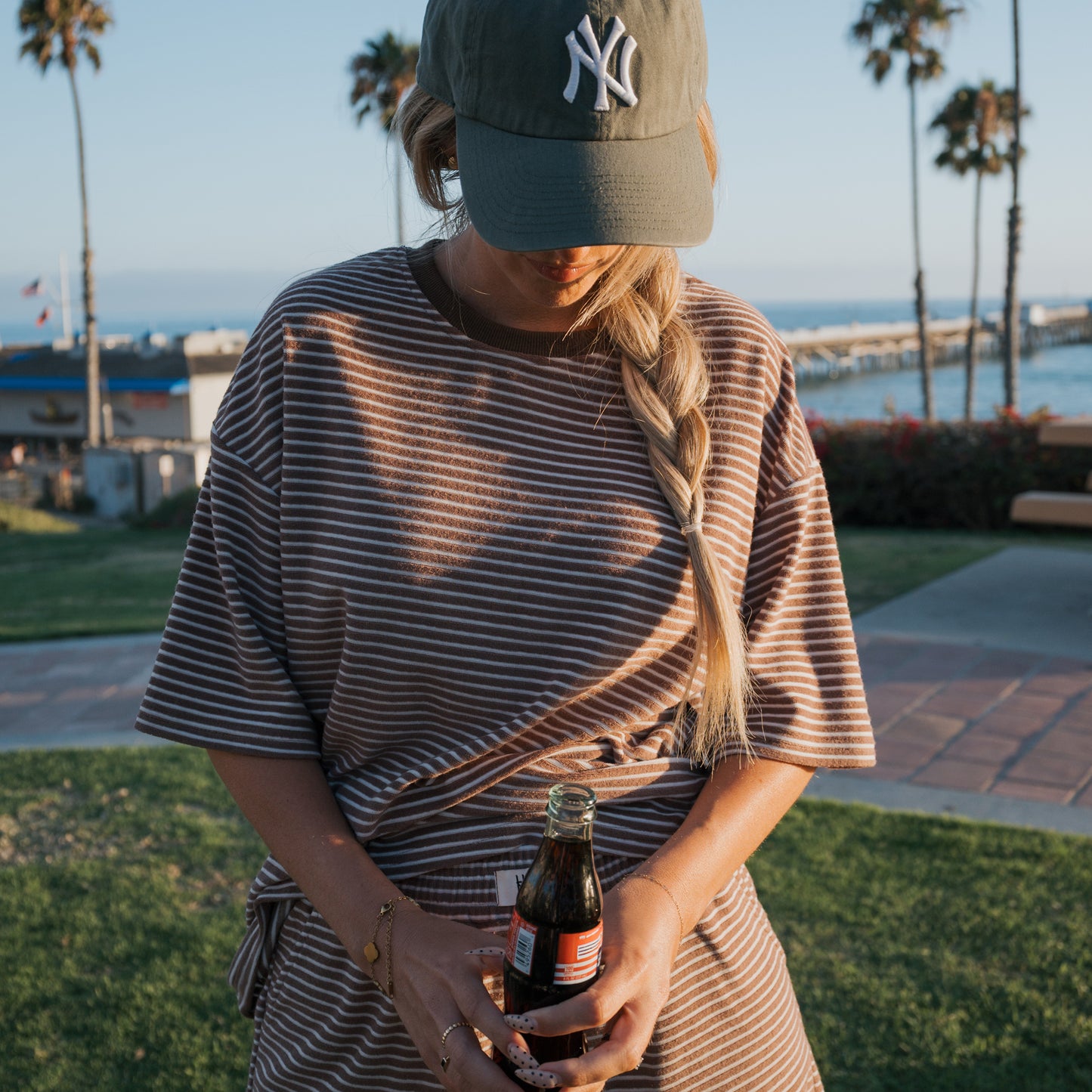 Wearing the Women's Terry Short Set in Cola Stripe by forever french baby, she stands outside by the ocean with palm trees and a pier behind her—perfect inspiration for relaxed loungewear or family beach outfits.