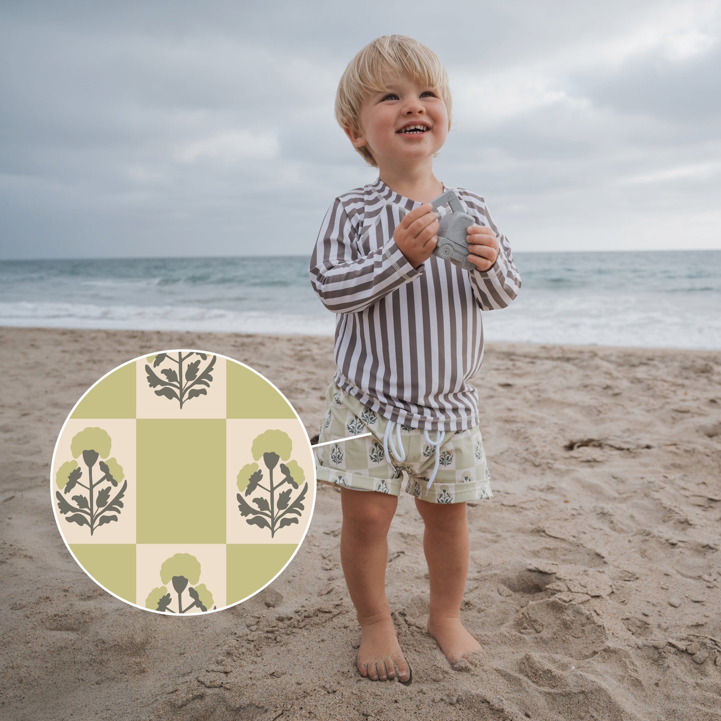 A toddler in a striped long-sleeve top and forever french baby Child Board Short | Marigold Check stands barefoot on the beach, smiling. An inset circle shows a close-up of the green and cream check pattern on these summer favorites.