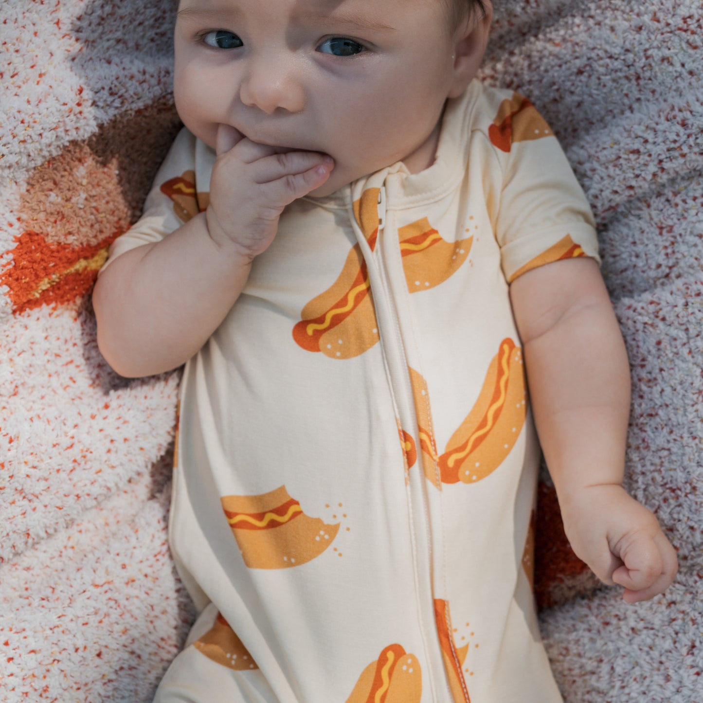 A baby lies on a textured blanket, wearing the forever french baby Shortie Bamboo Zip | Hot Dogs romper, with one hand in their mouth.