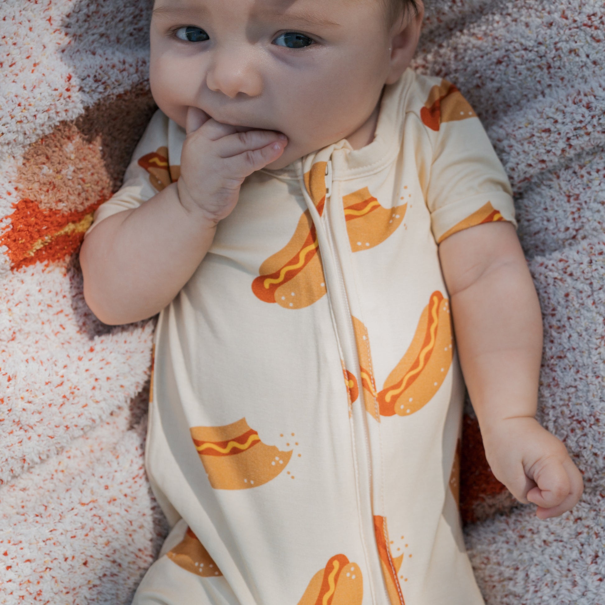 A baby lies on a textured blanket, wearing the forever french baby Shortie Bamboo Zip | Hot Dogs romper, with one hand in their mouth.