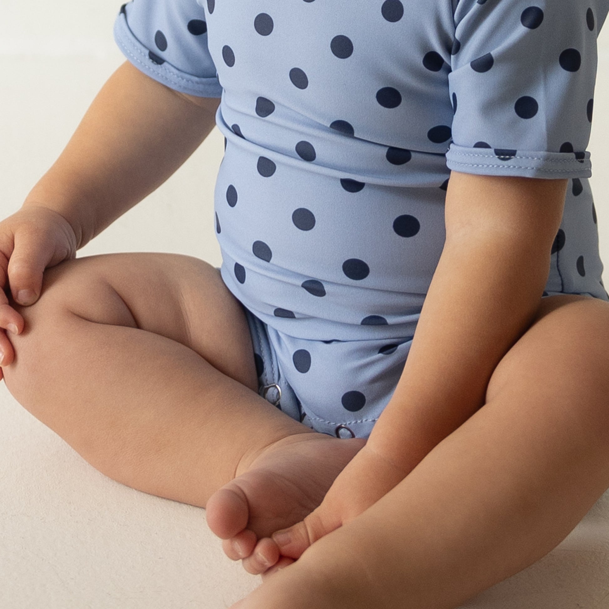 A baby, with only the lower half visible, sits cross-legged on a white surface wearing the forever french baby Child Shortie Swimsuit | Sully in light blue with dark blue polka dots.