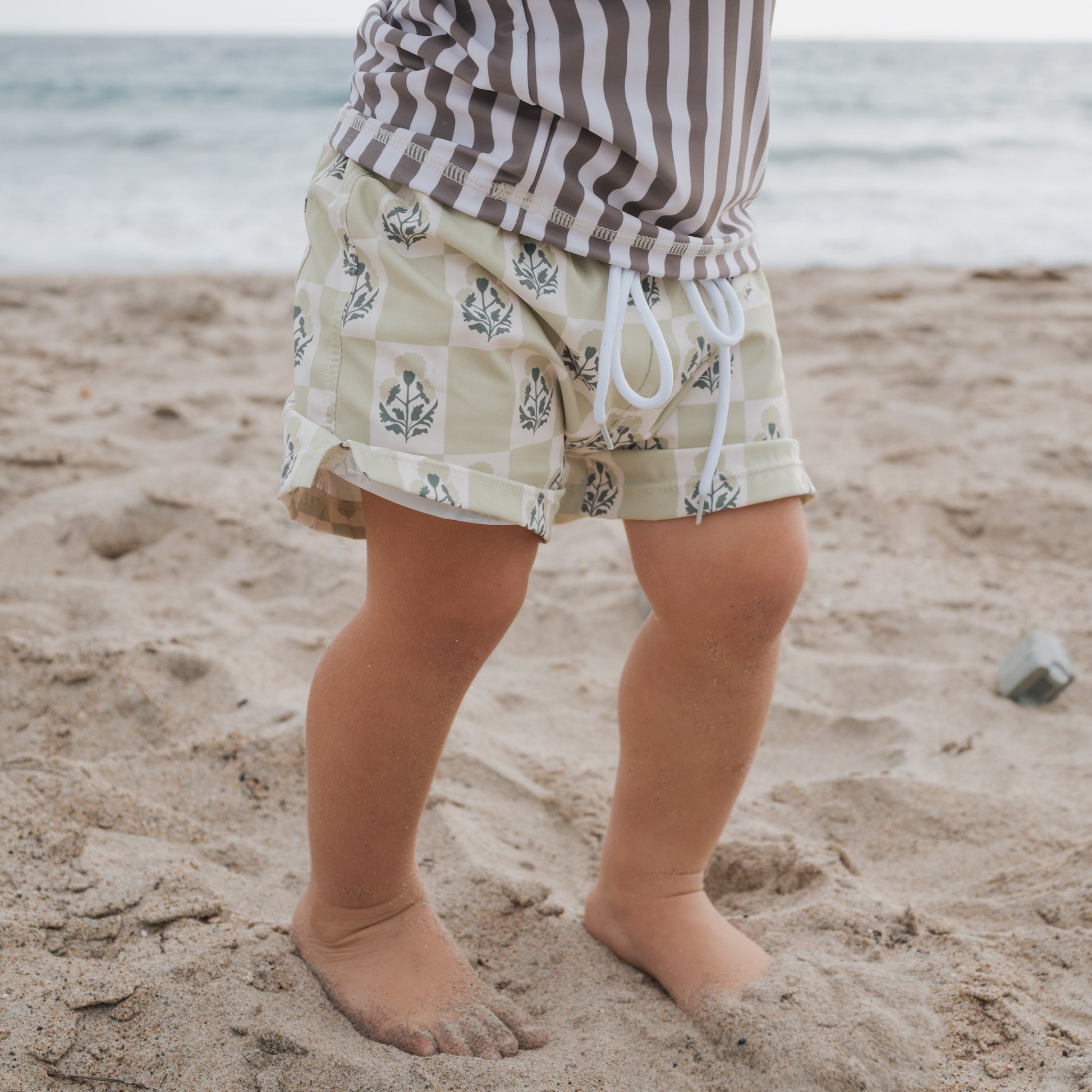 A small child stands barefoot on the sandy beach, wearing forever french baby's Child Board Short in Marigold Check. Ocean waves roll in behind, capturing a playful seaside moment in stylish kids activewear.