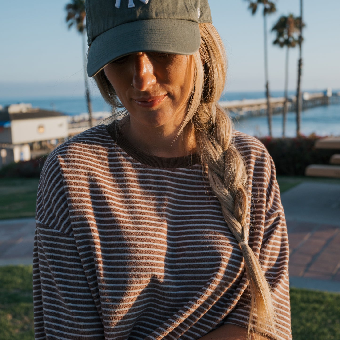 A woman in a green baseball cap and side braid stands by the beach, wearing the forever french baby Women's Terry Short Set in Cola Stripe. Palm trees, buildings, and a pier are bright in the background.