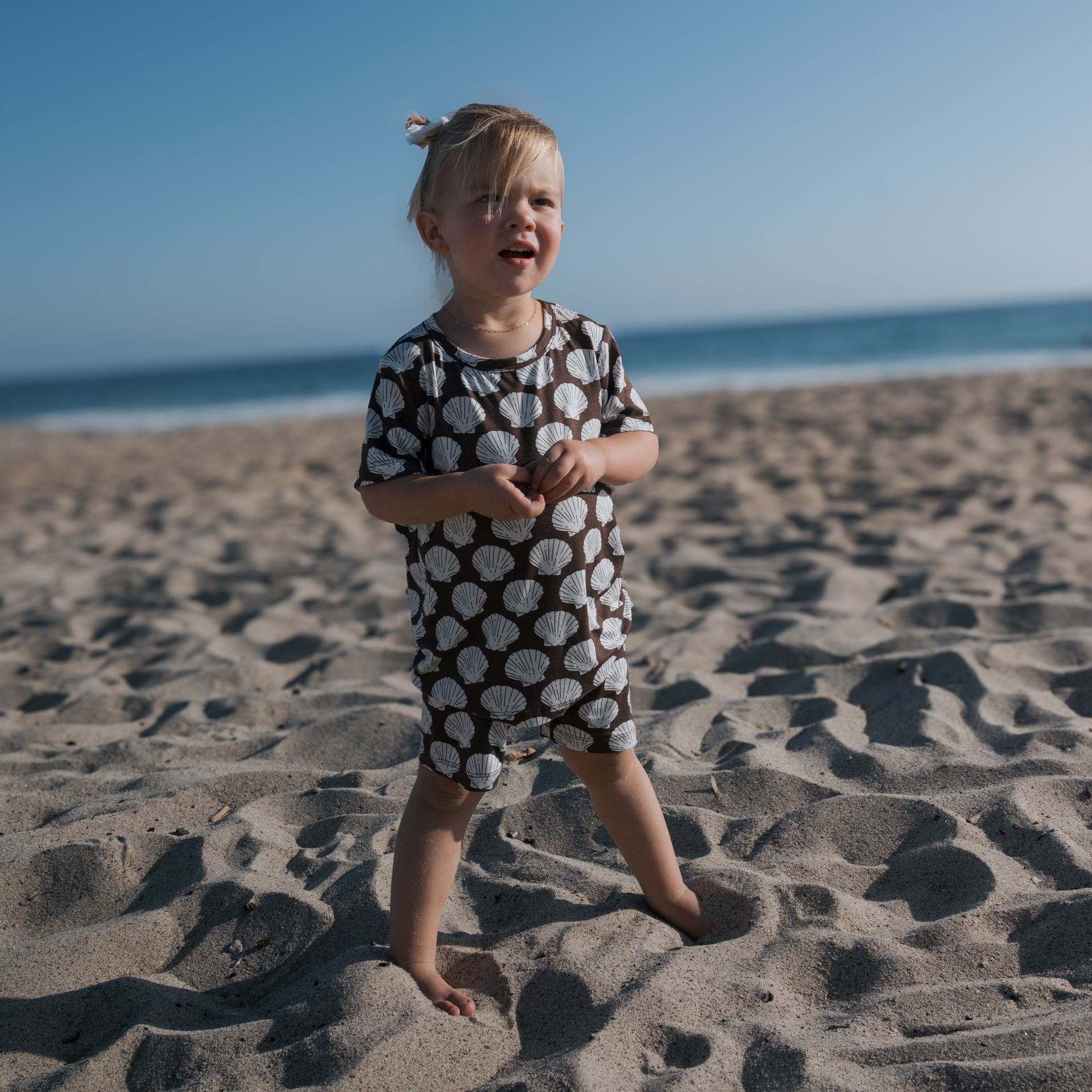 A young child smiles barefoot on the sand, wearing the forever french baby Bamboo Short Set | Seashells. The ocean and clear blue sky provide a beautiful backdrop.