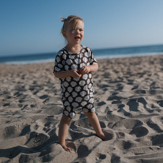 A young child smiles barefoot on the sand, wearing the forever french baby Bamboo Short Set | Seashells. The ocean and clear blue sky provide a beautiful backdrop.