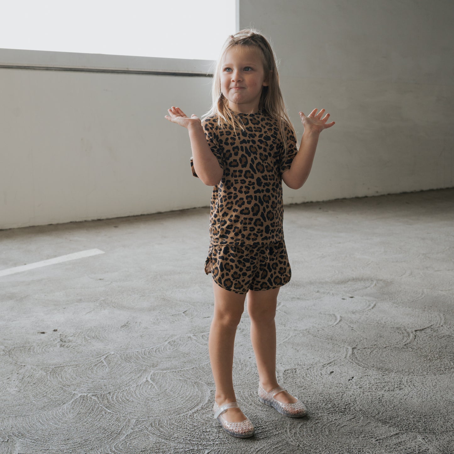 A young girl stands on a textured concrete floor wearing the forever french baby Girl's Bamboo Short Set | Leopard and translucent shoes, her hands raised with a playful expression.
