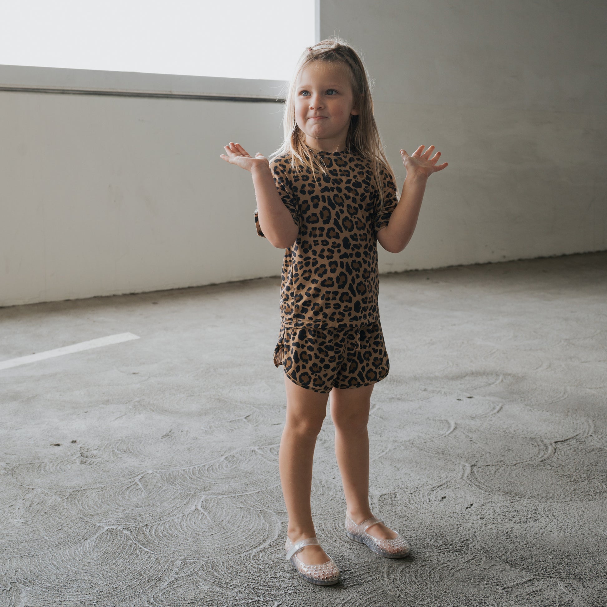 A young girl stands on a textured concrete floor wearing the forever french baby Girl's Bamboo Short Set | Leopard and translucent shoes, her hands raised with a playful expression.