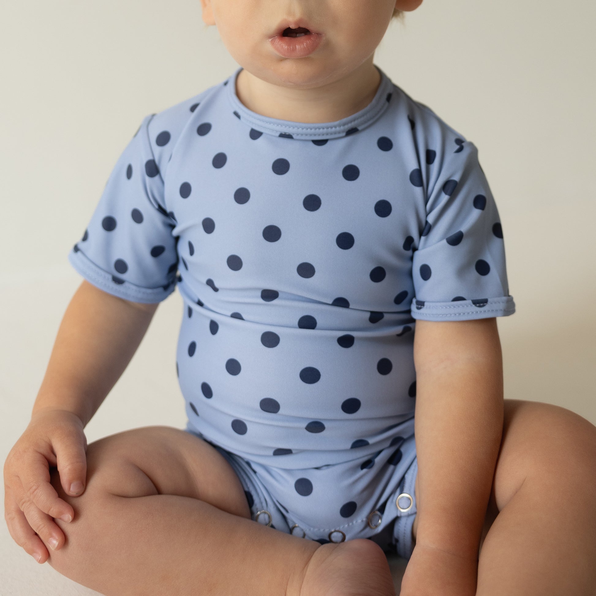 A baby sits cross-legged in the forever french baby Child Shortie Swimsuit | Sully, a light blue suit with dark blue polka dots. The baby's face is partially out of frame, highlighting the charm of this adorable swimwear.