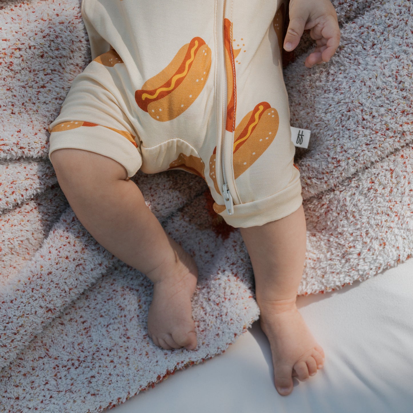 A baby wearing the forever french baby Shortie Bamboo Zip | Hot Dogs romper lies on a textured, light blanket. Only their legs, feet, and one hand are visible, highlighting the comfort of this hypoallergenic bamboo outfit.