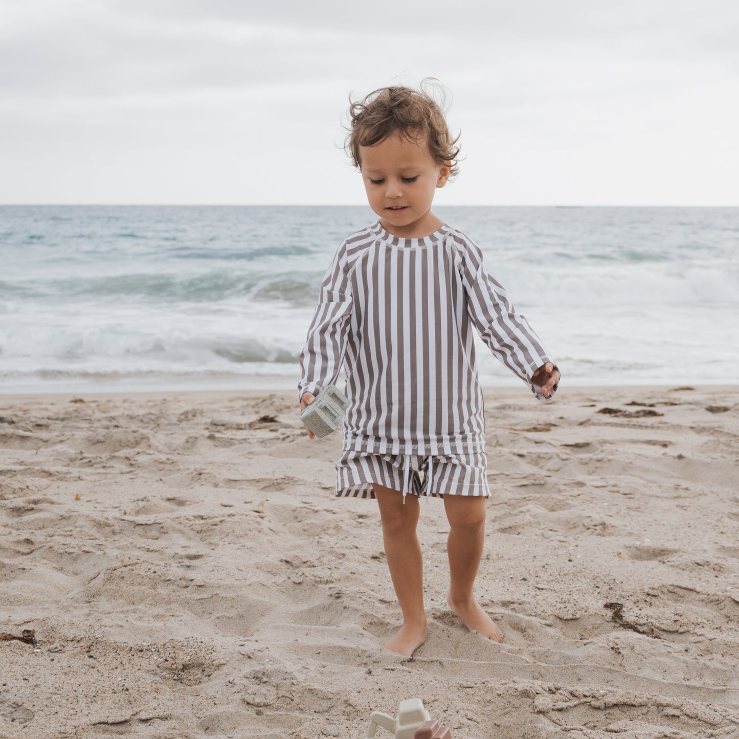 A young child in the forever french baby Child Board Short | Olive Stripe stands barefoot on a sandy beach near the ocean, holding a small object as waves crash in the background on a cloudy day.