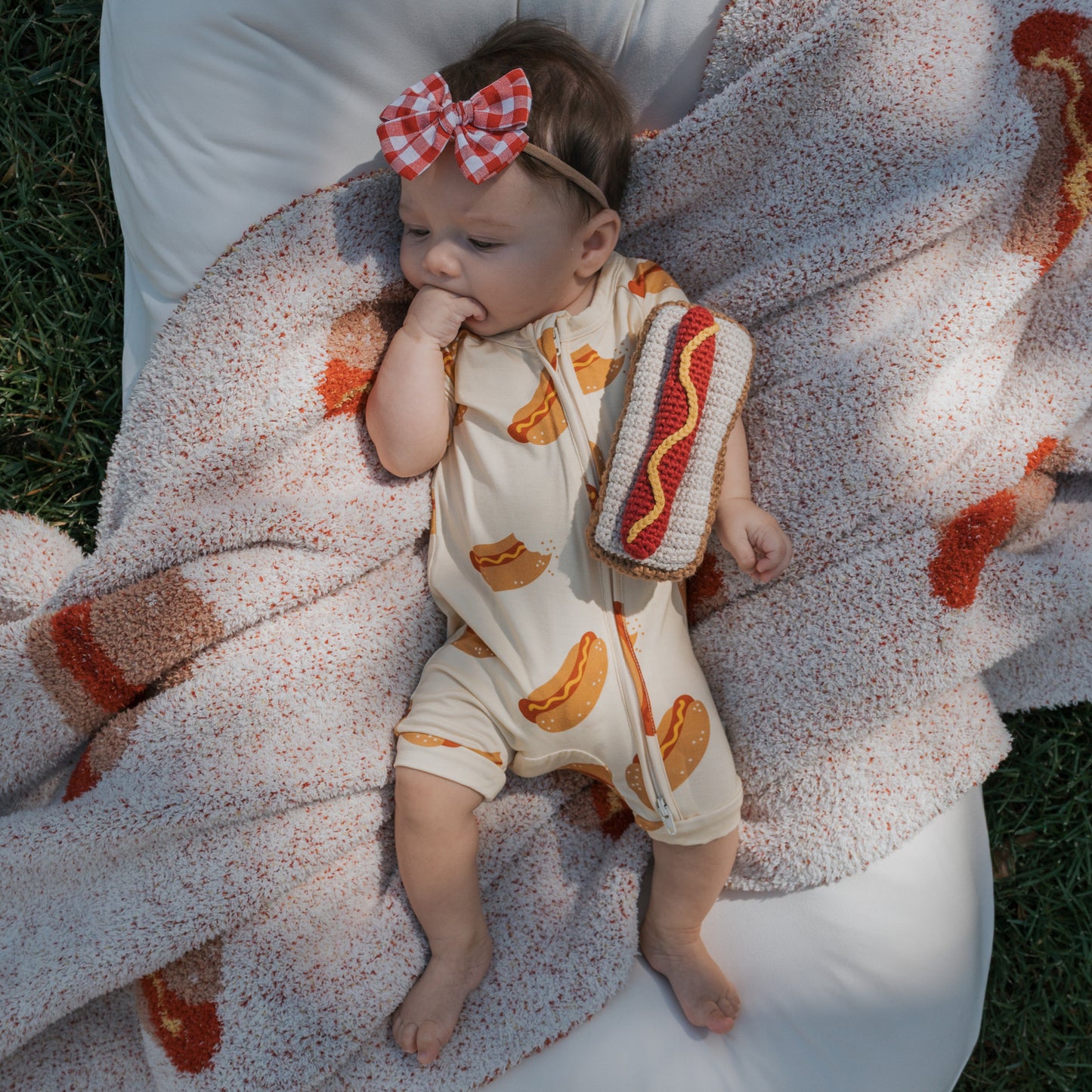 A baby in the forever french baby Shortie Bamboo Zip | Hot Dogs romper lies on a hot dog blanket, wearing a red plaid bow headband, holding a plush hot dog toy, and sucking their hand.