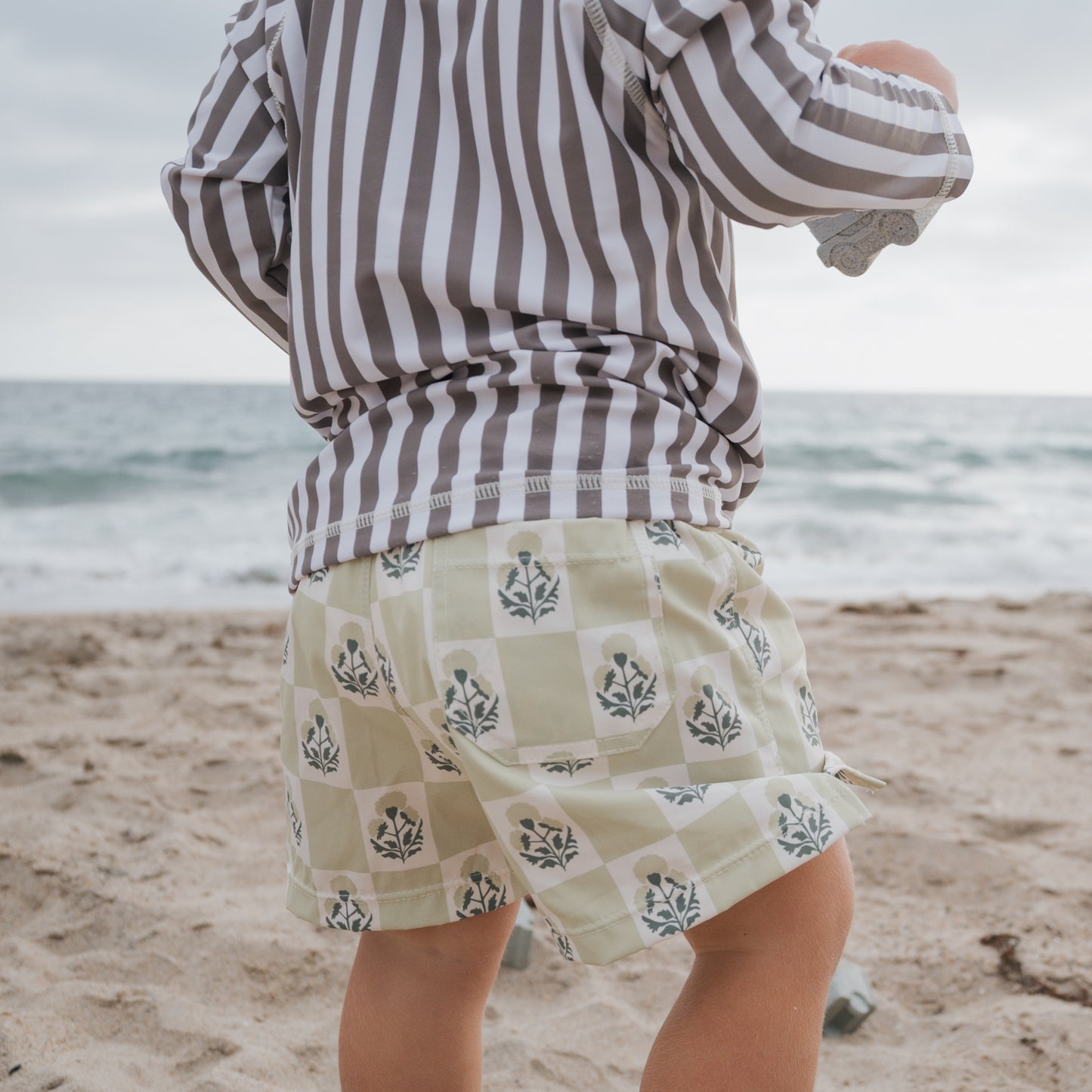 A young child in a striped long-sleeve shirt and forever french baby Child Board Short | Marigold Check stands on a sandy beach near the ocean, facing away from the camera with waves and a cloudy sky in the background.