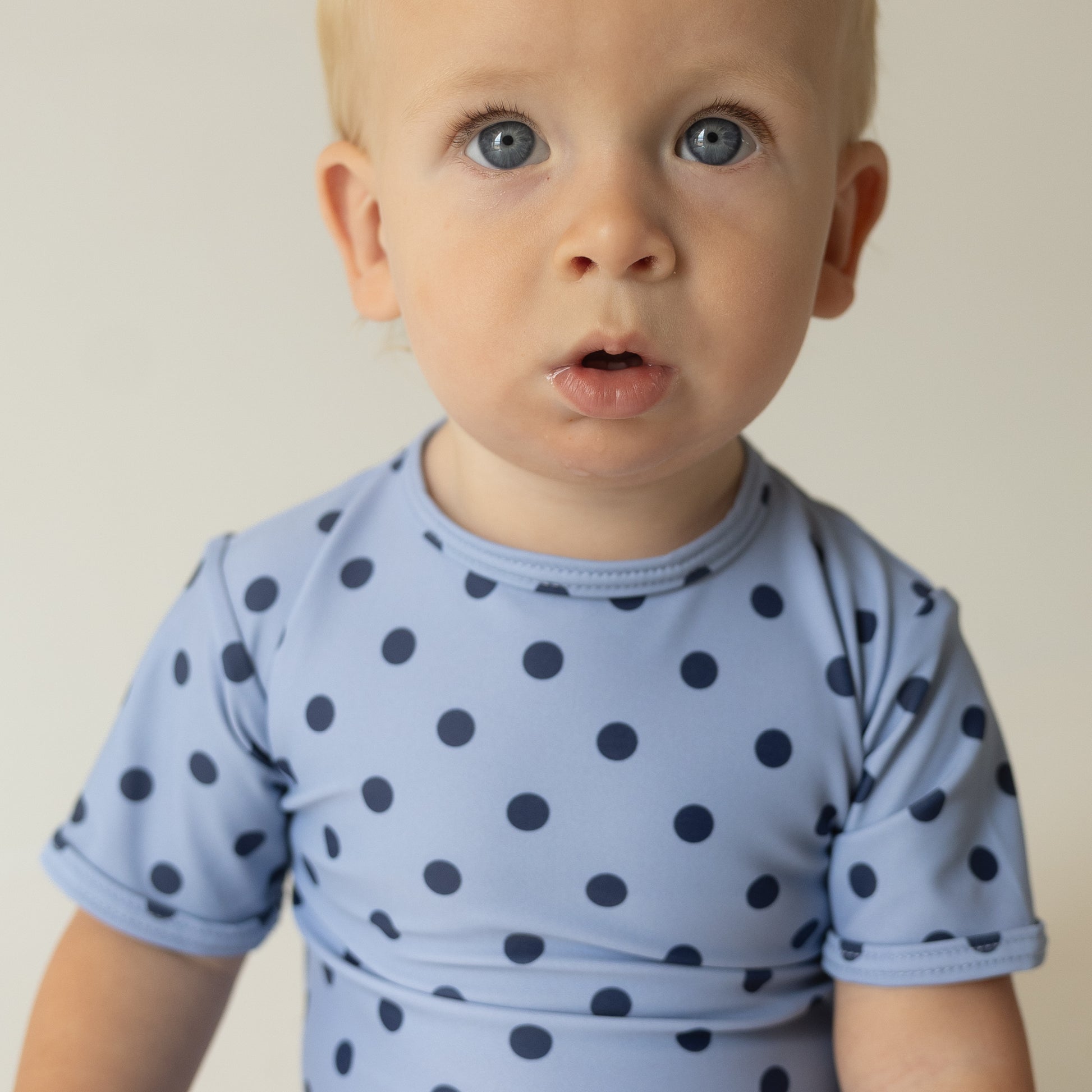 A toddler with blonde hair and blue eyes wears a light blue shirt with dark blue dots, capturing the playful style of the Child Shortie Swimsuit | Sully from forever french baby—perfect inspiration for your little one’s swimwear.