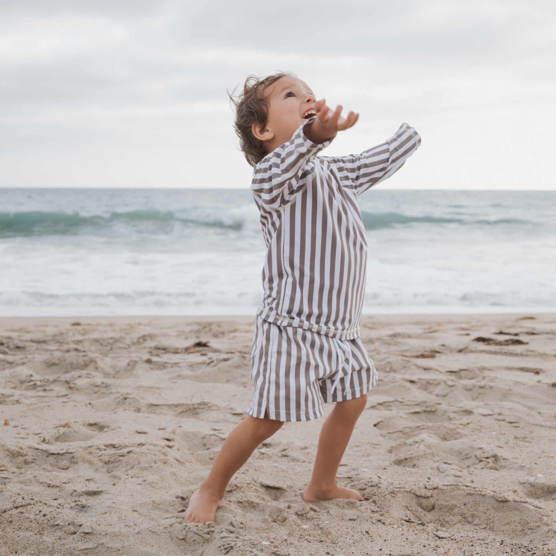 A child in forever french baby’s Olive Stripe Child Board Short smiles with arms outstretched on a sandy beach, wearing a long-sleeve striped shirt, barefoot with waves and a cloudy sky in the background.
